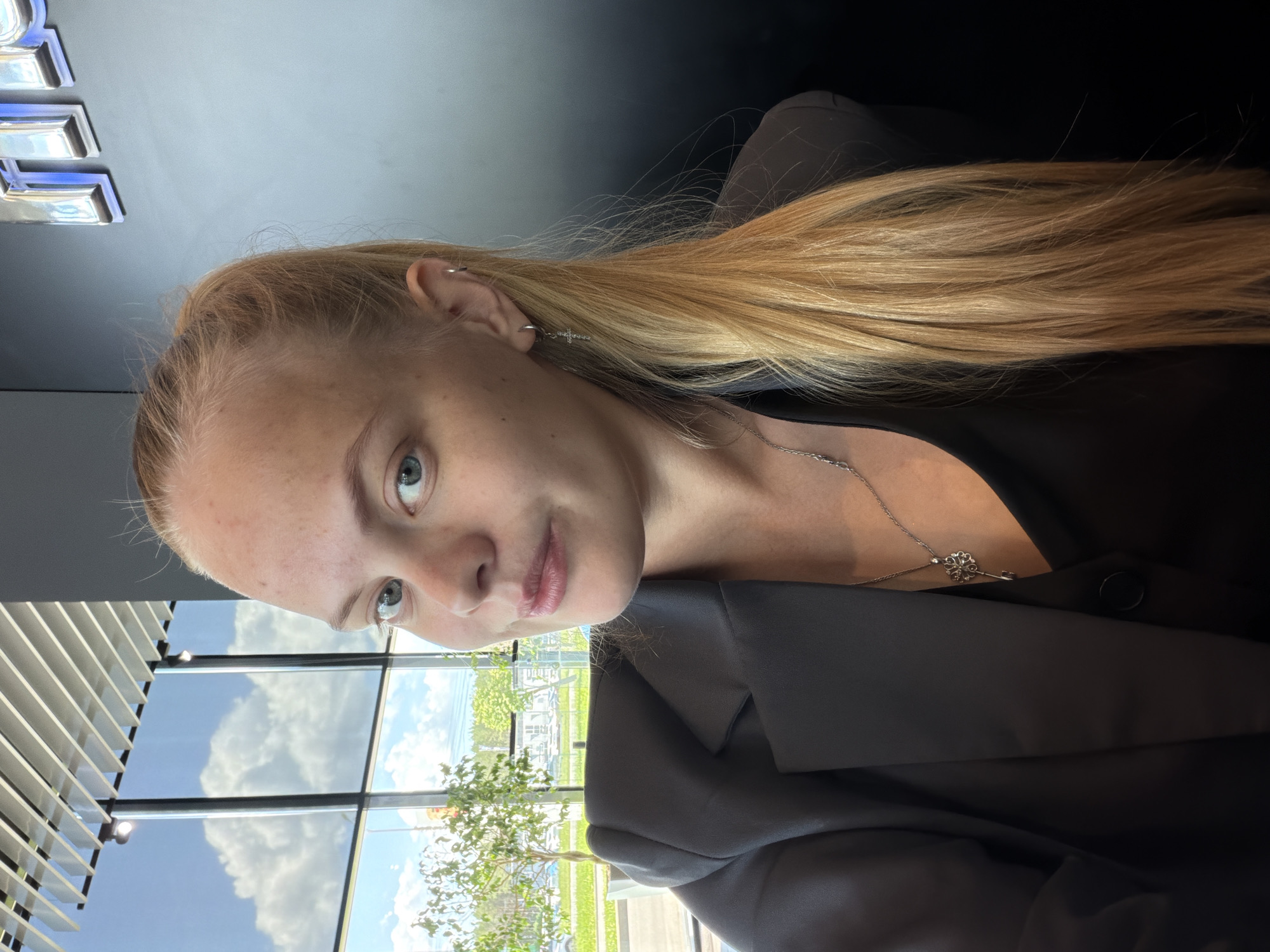 A blonde woman with her hair in a ponytail is sitting indoors, facing the camera. She is wearing a dark blazer over a dark top and a silver necklace with a key pendant. She has earrings in both ears, with one being a small hoop and the other a dangling cross. She is looking directly at the camera with a slight smile.
In the background, large windows show a view of trees, cars, and a cloudy blue sky. There is also a large, dark wall with a silver logo on it. The interior appears to be a modern building, possibly an office or a showroom, with a slatted ceiling and track lighting visible.
The image appears to have been taken in Izhevsk, Russia, given the context.