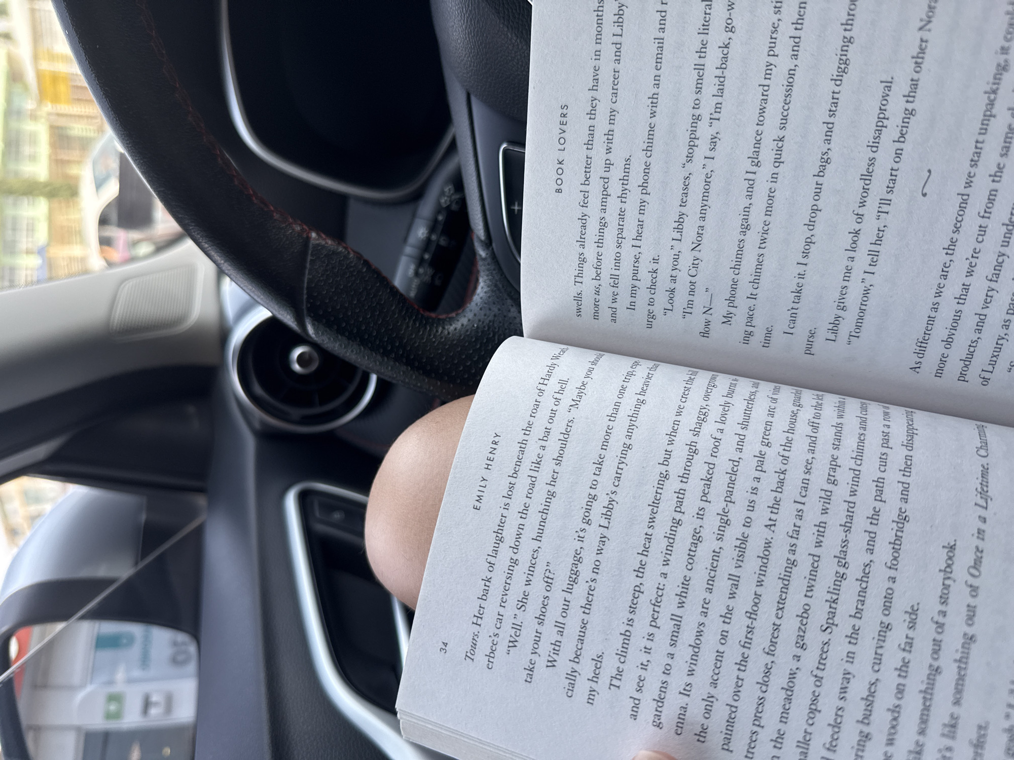 The image shows a book titled "Book Lovers" by Emily Henry, opened and resting on a person's arm inside a car. The steering wheel of the car is visible in the background. The car interior is dark, with glimpses of the exterior visible through the window. The book's pages are filled with text, and the pages are numbered as "34" on one side. The image's location in San Bernardino Tlaxcalancingo, Mexico is undetermined.