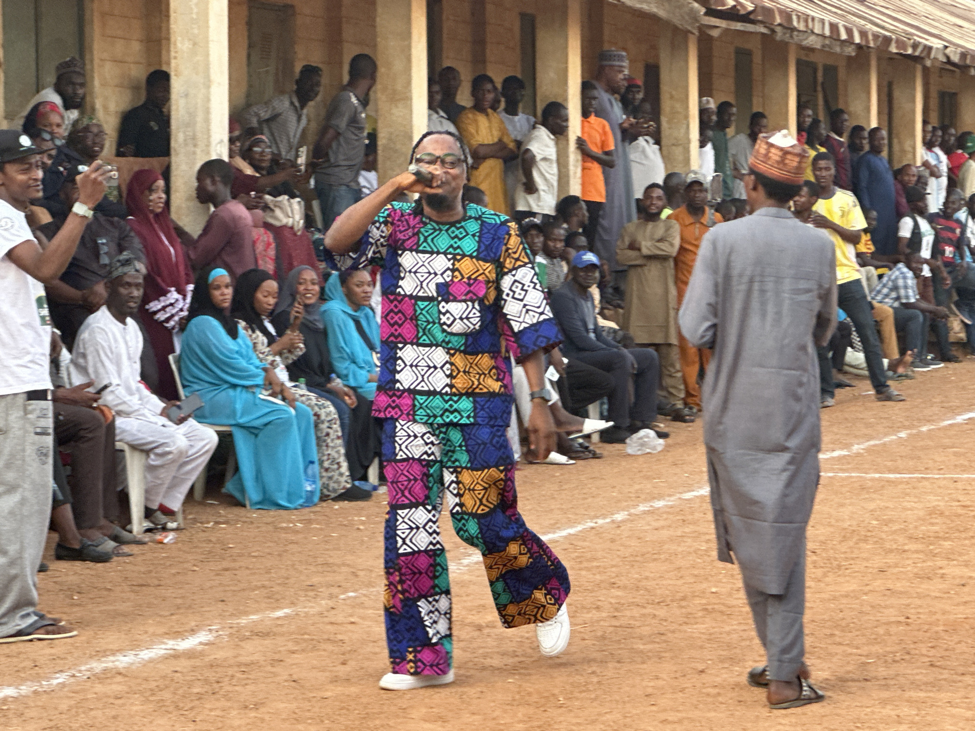 The image shows a gathering of people in an outdoor setting, possibly a cultural event or ceremony. A man in a colorful, patterned outfit is in the foreground, appearing to be speaking into a microphone or similar device held to his mouth. To his right, another man in a gray traditional outfit with a hat is walking in the opposite direction. A line of seated individuals is visible in the background, and numerous other people are standing or sitting, observing the scene. The ground is reddish-brown, suggesting a dirt or clay surface. The presence of buildings with pillars and a corrugated metal roof implies the location is in the city of Unknown, Unknown. The scene suggests a community event with a public address or performance occurring.