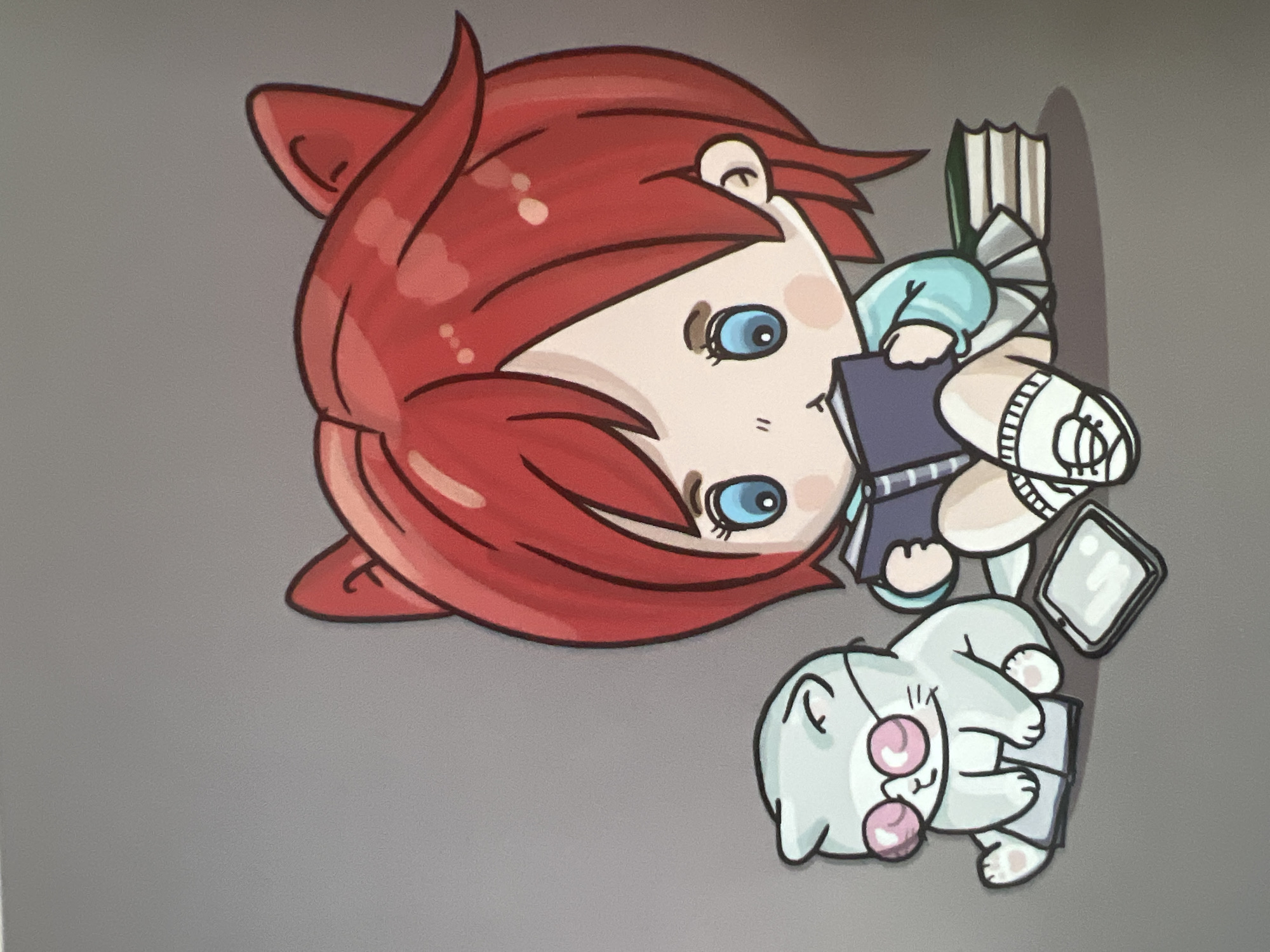 A digital illustration depicts a chibi-style anime girl with vibrant red hair and cat-like ears, sitting cross-legged and engrossed in reading a book. Beside her, a pale blue cat wearing pink-rimmed glasses also sits with its paws resting on an open book. Several stacks of books are placed near the girl, and a tablet-like device lies on the ground. The background is a plain, neutral gray. There is no indication of any specific location in Krasnodar, Russia, or any urban environment. The scene is entirely fictional and cartoonish.