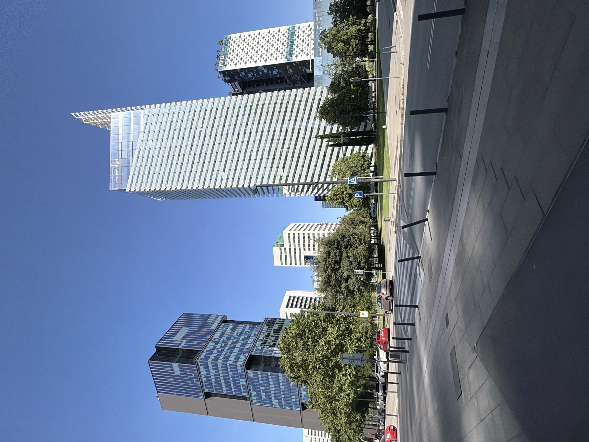 The image captures a cityscape in L'Hospitalet de Llobregat, Spain, featuring several tall buildings against a clear blue sky. The buildings vary in design; one has a diagonally striped facade, another a grid-like pattern, and others with a more traditional office building appearance. Trees line the street, partially obscuring some of the buildings. A street with crosswalks and lamp posts is visible, with parked cars along the side. The sidewalk takes up a significant portion of the foreground.