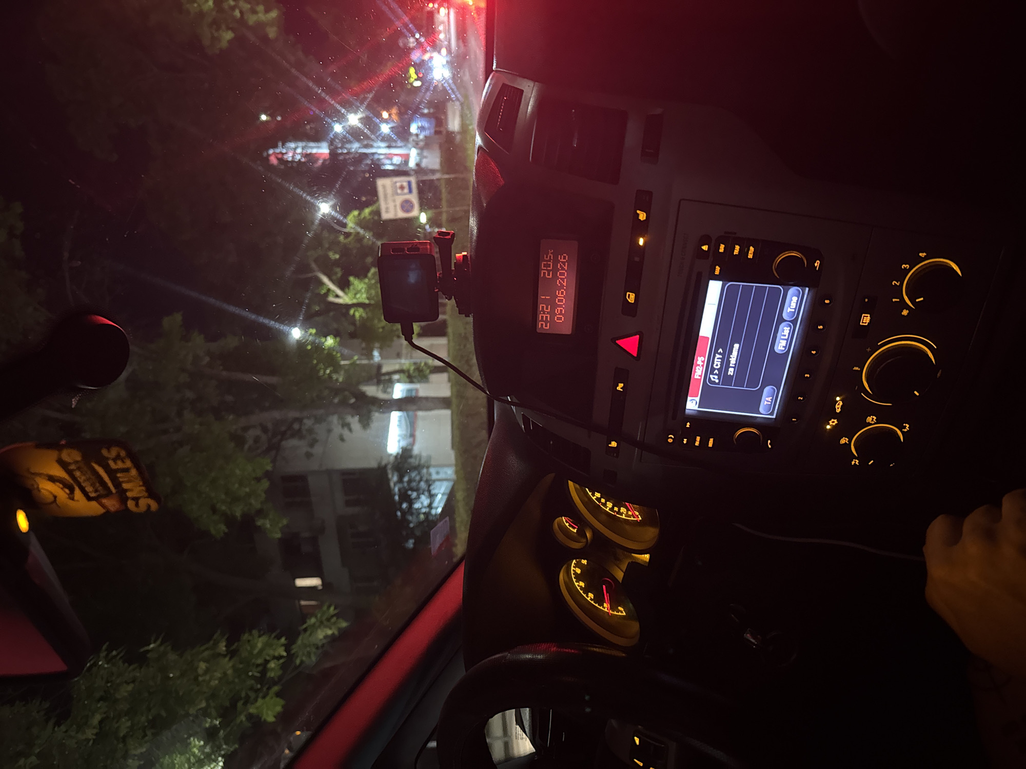 The image shows the interior of a car at night, as seen from the driver's perspective. The dashboard is illuminated with orange and red lights. A digital display shows the time as 23:21 and the date as 09.06.2026. A small black camera is mounted on top of the dashboard. The car's radio display shows "FM2-PS" at the top, with text in a foreign language below, including "> CITY >" and "za reklama". The steering wheel is visible on the left. Outside the car, through the windshield and side windows, streetlights and building lights are visible, suggesting an urban environment. Trees and foliage are also present. A hand is visible in the lower left corner of the frame, appearing to be on the gear shift.