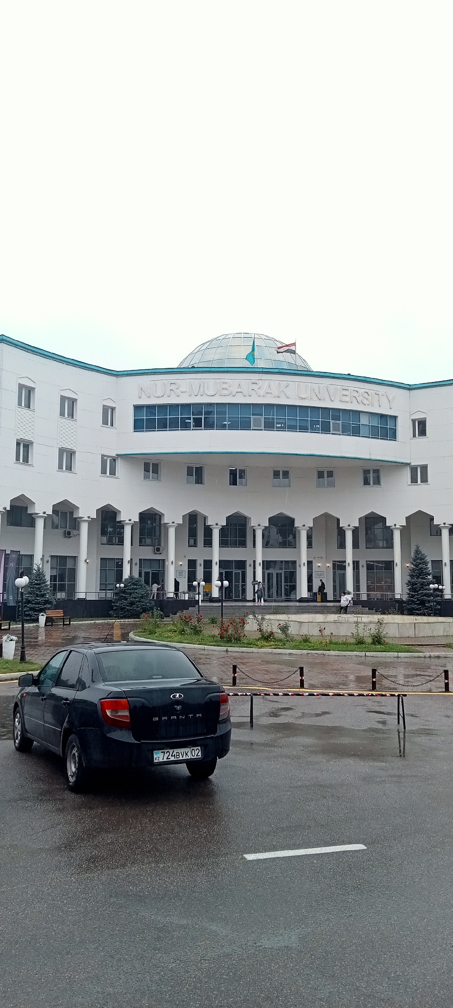 The image depicts the front exterior of Nur-Mubarak University in Almaty, Kazakhstan. The building is a modern, white, multi-story structure with a prominent glass dome on its roof, topped with the flags of Kazakhstan and Egypt. The building's facade features a colonnade of white pillars leading to arched entrances. In front of the building, a black sedan is parked on a wet asphalt surface, suggesting recent rain. A black license plate with the inscription "KZ 724BVK 02" is visible on the car. Several people are visible near the building's entrance, some walking and others standing. The surrounding environment includes well-maintained landscaping with trees and flower beds. The sky is overcast.