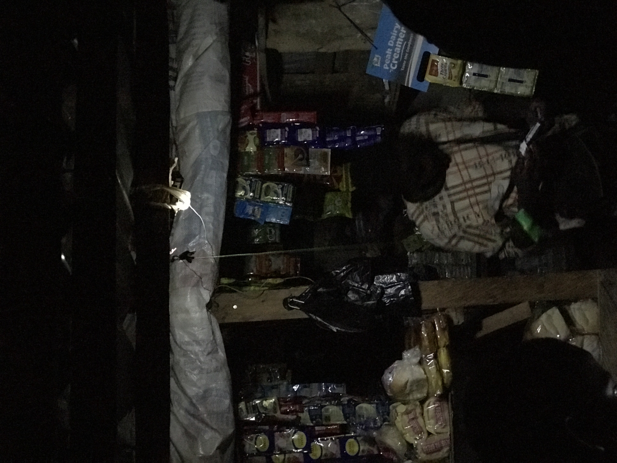 The image depicts a dimly lit, enclosed space appearing to be a small shop or storage area in Abuja, Nigeria. Shelves constructed of wood hold stacks of packaged goods. Visible products include Peak Dairy Creamer boxes and individual packets of Three Crowns brand milk. Also seen are wrapped baked goods (likely bread). A large, shrouded object, possibly a rolled-up tarp or fabric, leans against a vertical support structure. The overall setting suggests a basic, low-income retail environment.