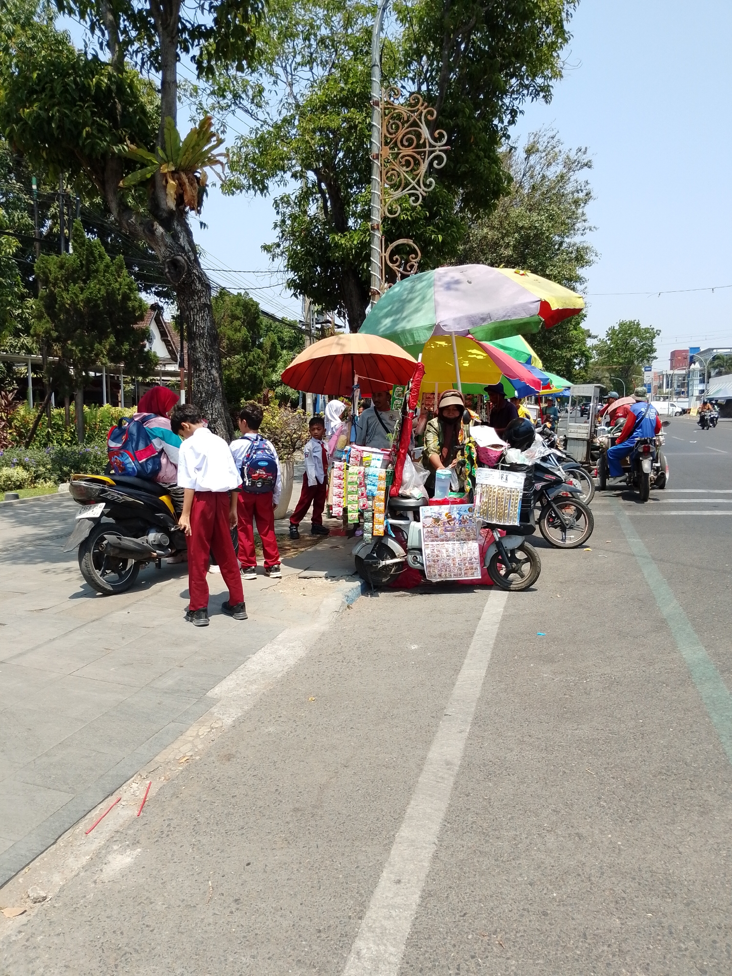 In the city of Pasuruan, Indonesia, an outdoor scene depicts a bustling street with several people and vendors. In the foreground, a scooter with a rider is parked, its passenger side facing the camera. To the left of the scooter, two young boys in school uniforms, red pants, and white shirts stand on a sidewalk. One boy, facing away from the camera, is bent over. Another boy, wearing a white shirt and red pants, stands with his back to the camera. The sidewalk is paved with grey stones.
Further down the sidewalk, a street vendor operates from a cart adorned with colorful umbrellas. The cart displays various goods, including packaged items and what appear to be lottery tickets. A woman with a head covering sits behind the cart, attending to customers. Two children in school uniforms stand near the vendor, one in a white shirt and red pants and the other in a white shirt, red pants, and a blue backpack. Another child in a white shirt and red pants is walking away from the vendor.
Several other vendors and their carts are visible further down the street, also with colorful umbrellas providing shade. Motorcycles are parked alongside the carts. In the background, buildings and trees line the street, and a clear blue sky indicates a sunny day. A blue motorcycle with a rider in a blue outfit and a red helmet is visible on the road to the right. The road surface is asphalt, with white lines marking lanes. Power lines are strung across the scene, and a decorative lamppost stands near the vendors.