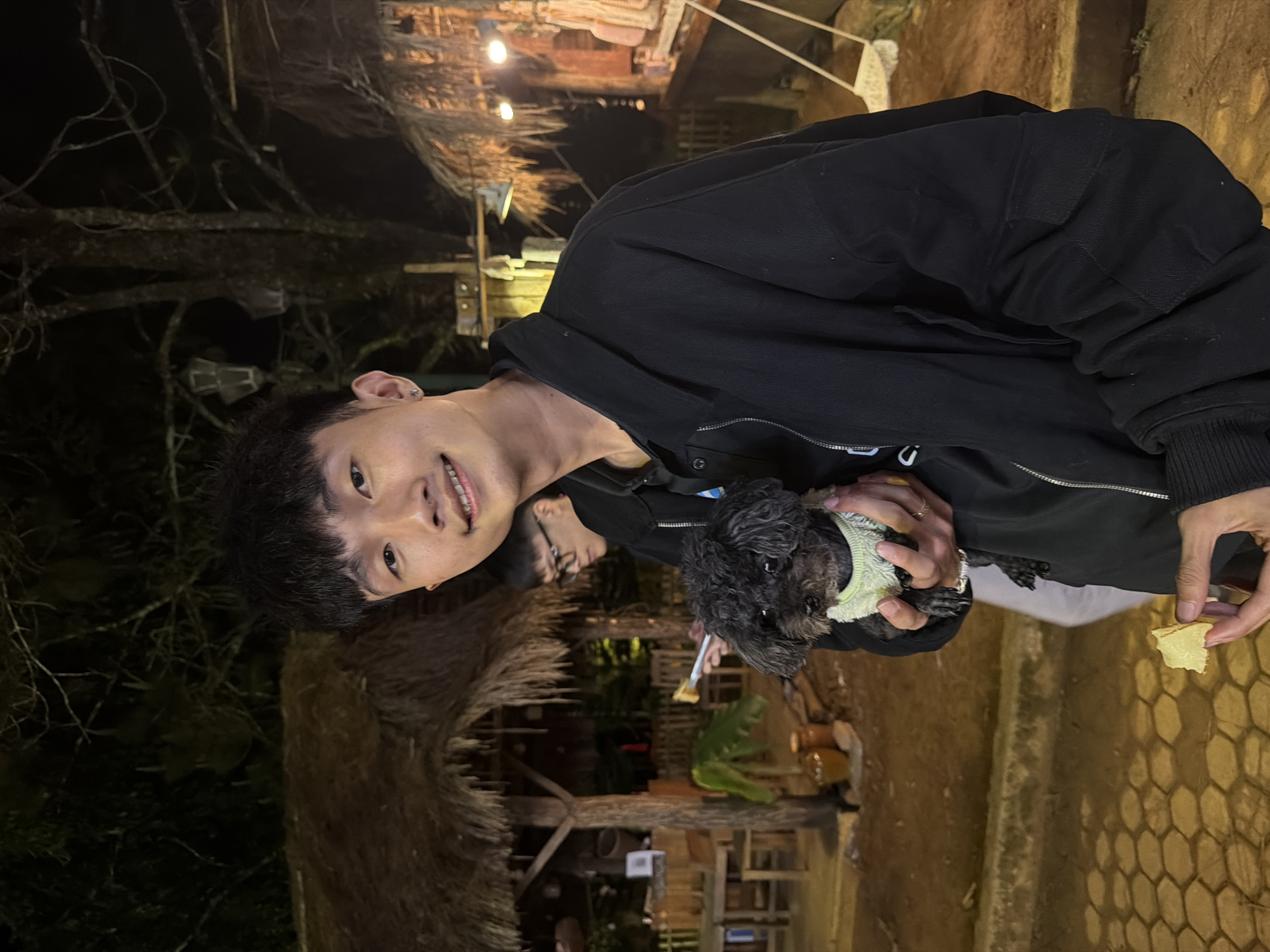 A young Asian man is captured in a nighttime outdoor setting, holding a small, black, fluffy dog. He is smiling warmly at the camera, wearing a black jacket, and has short dark hair. The dog, adorned in a light-colored sweater or vest, is held close to the man's chest. The man appears to be offering a small piece of light-colored food, possibly a cracker, to the dog, which looks up expectantly.
In the background, a second person is partially visible, wearing glasses. The scene is dimly lit by string lights and other artificial illumination, casting a warm glow on rustic wooden structures and thatched roofs, suggesting an outdoor cafe, resort, or homestay. Bare tree branches are visible against the dark night sky. The location context of Măng Đen, Vietnam, aligns with the natural and somewhat rustic aesthetic of the background. The man's joyful expression indicates a pleasant and affectionate interaction with the dog. No text is visible in the image.
