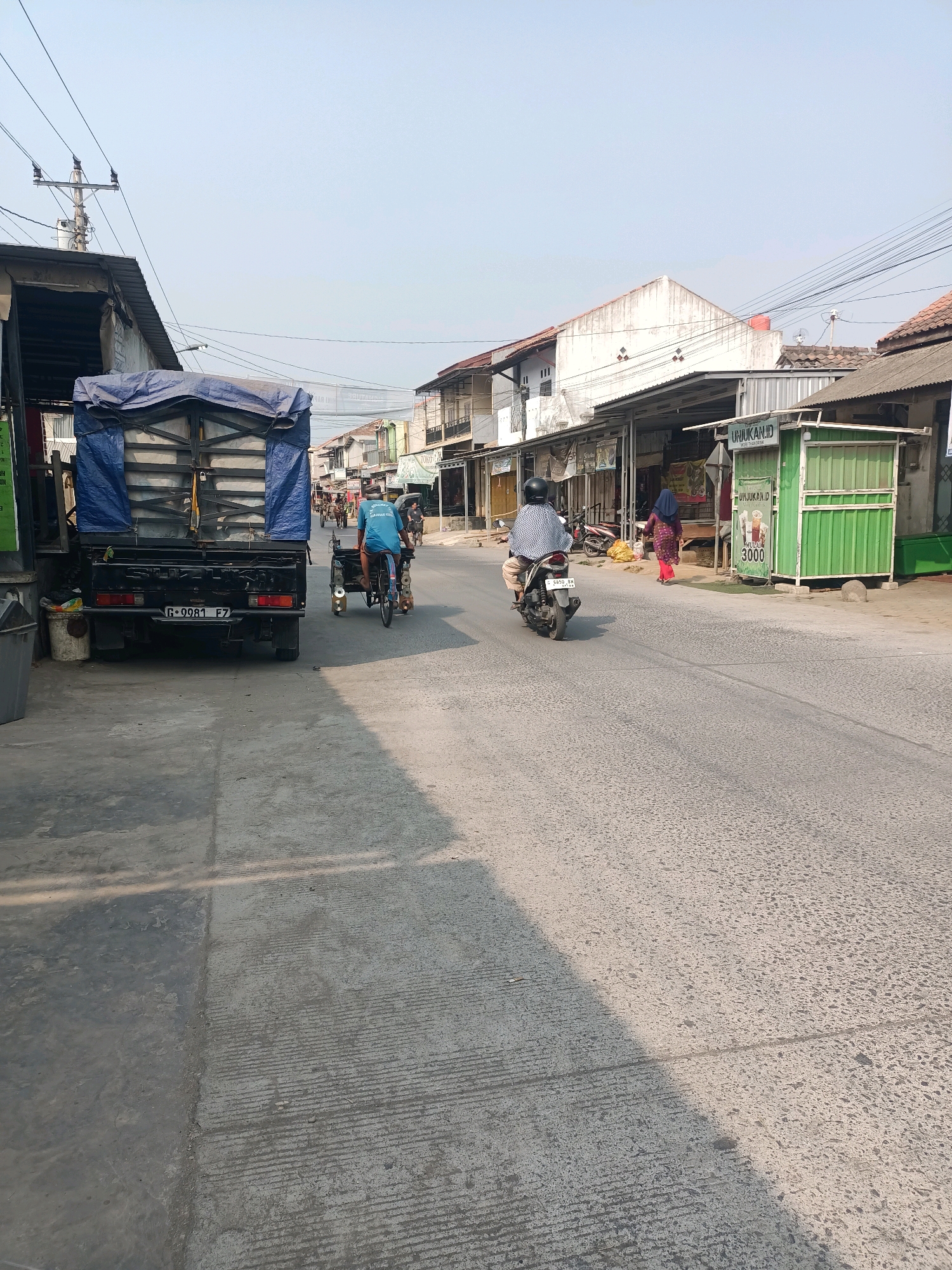 The image depicts a street scene in Tembok Banjaran, Indonesia. On the left, a black pickup truck with a blue tarp covering its cargo bed is parked. A garbage can is visible near the truck. In the middle of the street, a person wearing a blue shirt is riding a bicycle with a small trailer attached. Further down the street, a person on a motorcycle is riding away from the camera. To the right, a row of buildings with storefronts lines the street. One storefront is painted green and has a sign with text and an image of a drink. Another building to its left has a white facade. The sky above is clear and pale. Power lines are visible crisscrossing the sky. The road surface is asphalt with visible wear and texture.