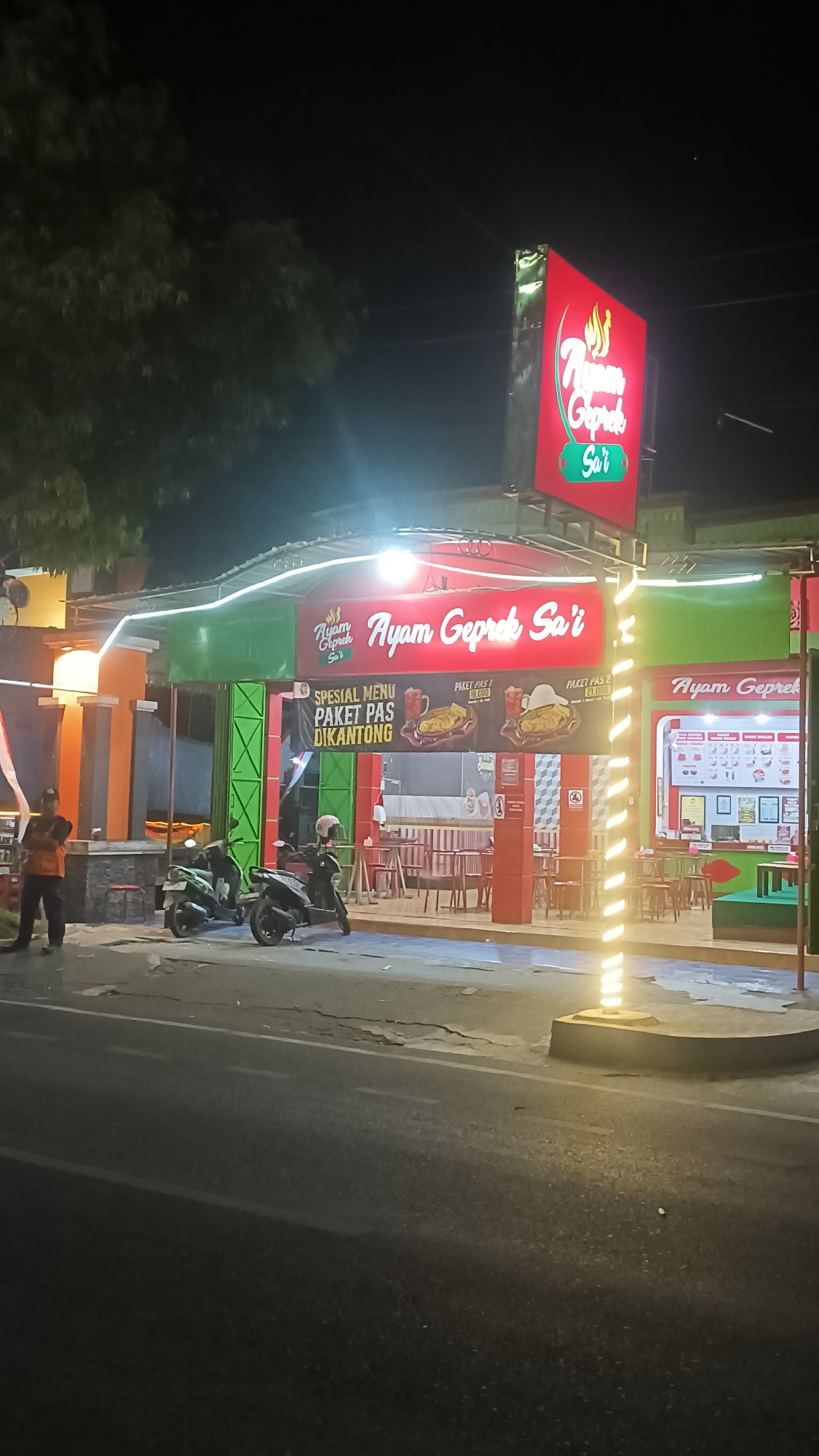 A night-time exterior shot captures a brightly lit restaurant called "Ayam Geprek Sa'i" in Sragen, Indonesia. The restaurant features a prominent red and green facade with a large, illuminated sign that reads "Ayam Geprek Sa'i" in white lettering. To the left, a man in an orange uniform stands near the entrance. Two motorcycles are parked in front of the restaurant. Banners displaying food items and prices are visible on the wall. The street in front of the establishment is paved and appears somewhat cracked. Trees can be seen in the background.