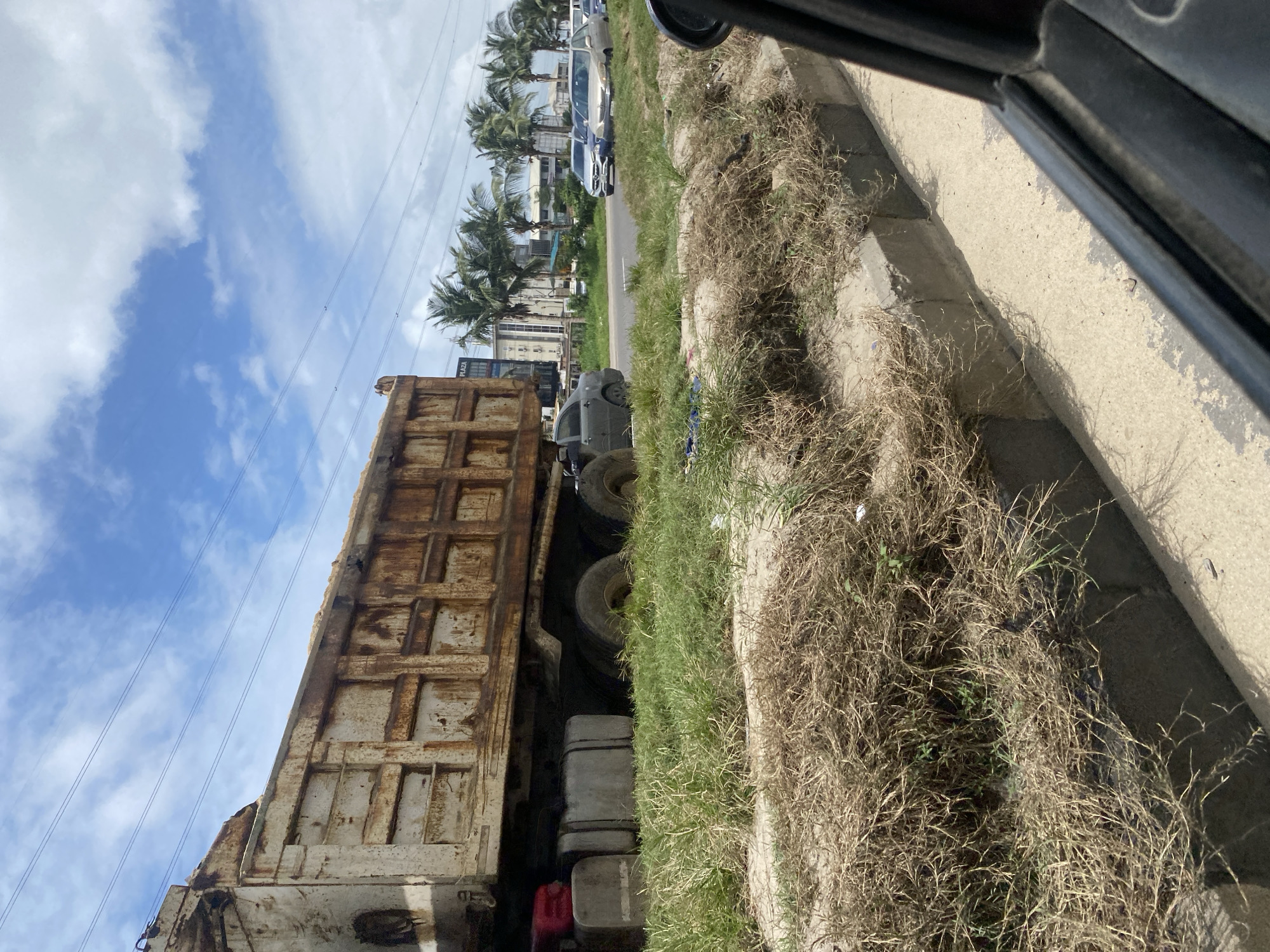 The image depicts a side view of a large, rusty dump truck parked on the side of a road in Lekki Peninsula II, Nigeria. The truck bed is filled with a light-colored granular material, likely sand or gravel. In front of the truck, several gray fuel tanks are visible. To the right of the dump truck, a dark-colored sedan is parked. Further down the road, a line of cars, including a silver SUV and a blue sedan, are visible. The background features tall palm trees and modern buildings, suggesting an urban or semi-urban environment. The sky is blue with scattered white clouds and overhead power lines. The foreground shows a grassy embankment with dry, straw-like vegetation and a concrete curb separating it from the road. The image is taken from within a vehicle, as indicated by the partial view of a black car interior on the right side of the frame.
