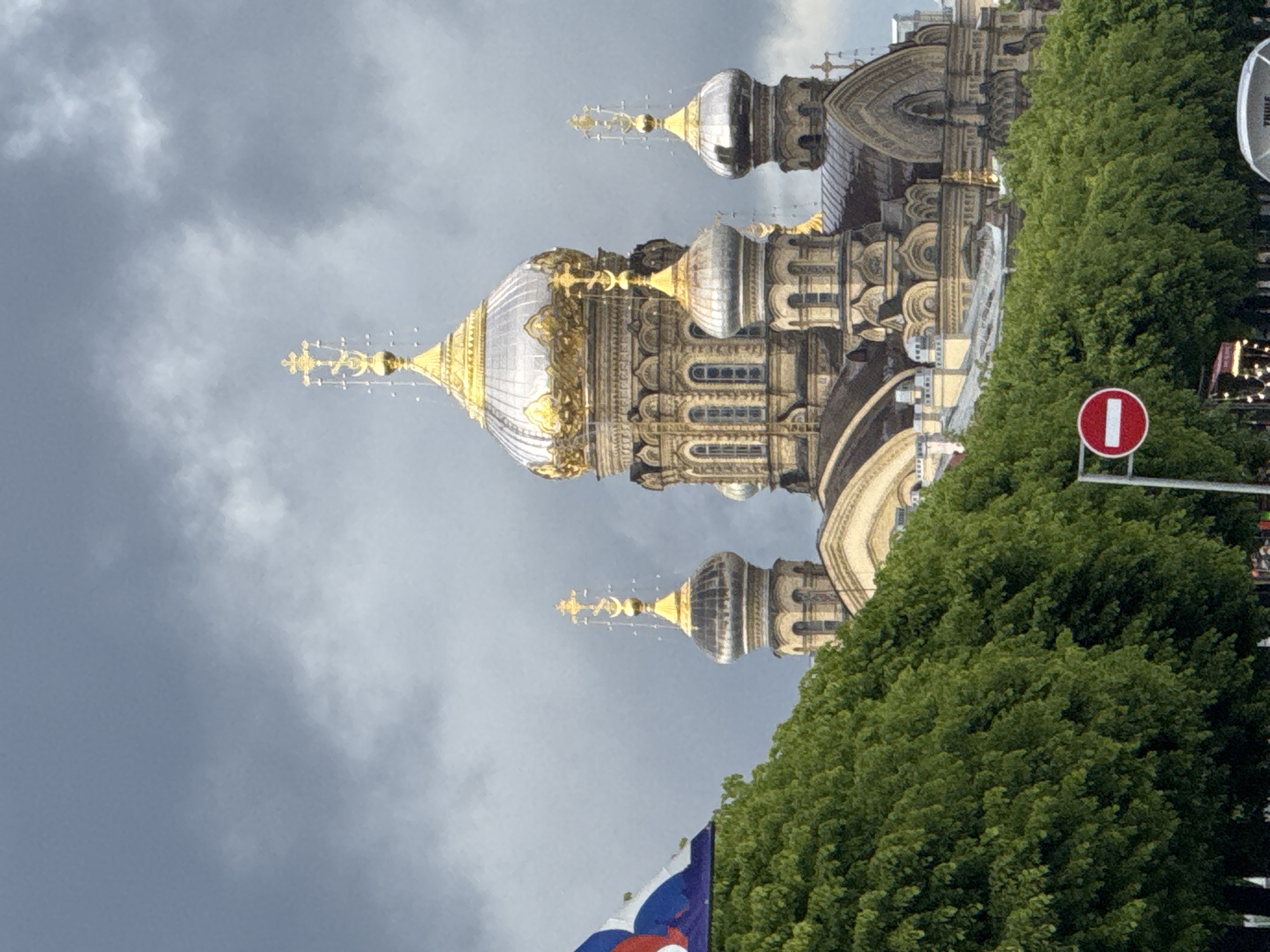 The image shows a Russian Orthodox church in an urban setting under an overcast sky. The building is ornate, with multiple domes, the largest of which is silver with gilded detailing. A row of green trees partially obscures the lower part of the church. A "do not enter" sign is visible to the right of the trees. A Russian flag is visible in the lower left corner. The location is unknown.