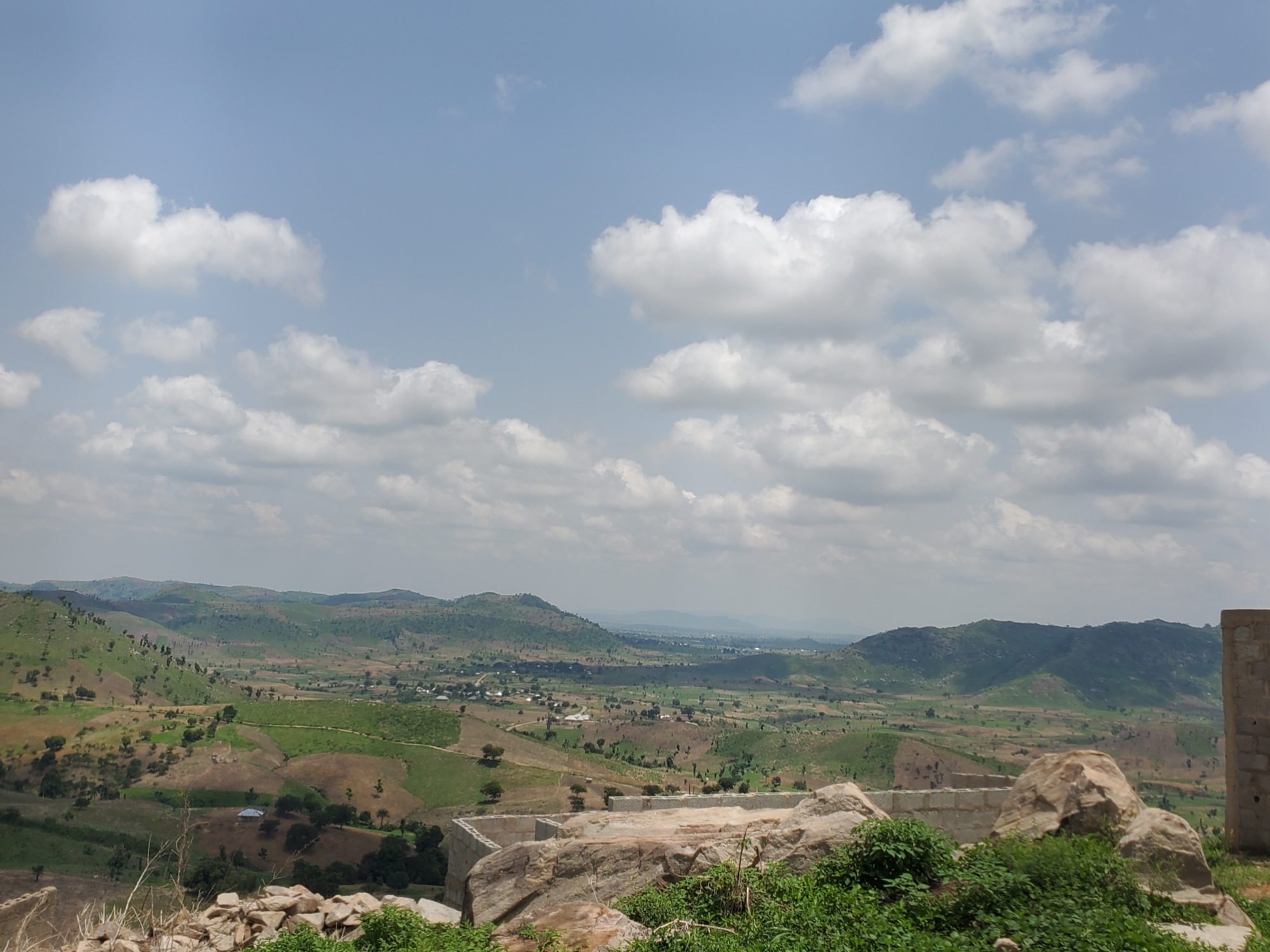 An eye-level, wide-angle, outdoor shot captures a vast, verdant landscape under a partially cloudy sky. The foreground is dominated by rugged, grey and tan rocks and a pile of rough-hewn stones, interspersed with vibrant green foliage and dry, tan grasses. Beyond this natural foreground, the terrain descends into rolling hills and valleys, a patchwork of green fields, brown cultivated land, and scattered trees. In the middle ground, a small cluster of buildings with light-colored roofs is visible, nestled among the hills. Further in the distance, the landscape continues with more undulating hills, fading into a hazy horizon. The sky is a pale blue, dotted with fluffy white cumulus clouds, some of which are tinged with grey, suggesting potential for light precipitation. A partially constructed building, made of concrete blocks, stands on the far right of the image, its rough texture contrasting with the natural elements. The overall scene depicts a rural, possibly developing, area with a natural, untamed beauty.