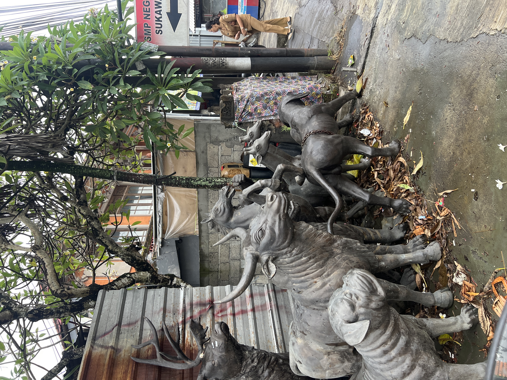 The image depicts an outdoor scene in Sukawati, Indonesia. A stack of weathered, grayish-bronze animal sculptures, including bulls, deer, and what appear to be goats or horses, sit on a wet, gray concrete surface covered with fallen leaves and debris. A tree with dense foliage is visible to the left, obscuring part of the building behind. A sign indicates "SMP NEGE... SUKAWA..." with a downward arrow. In the background, a woman in a brown uniform bends over near what appears to be a stack of pipes. The background architecture appears to be a mix of industrial and residential buildings. The overall scene appears to be a street or sidewalk in an urban environment.