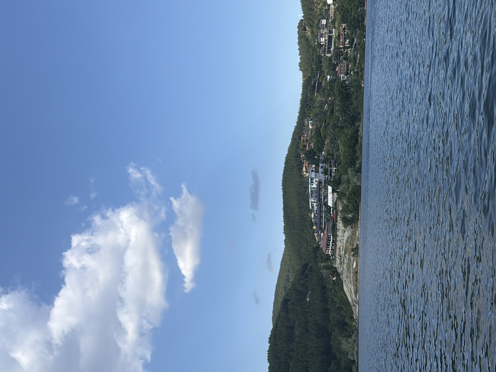 The image shows a wide, calm lake in the foreground. In the background, a densely wooded hillside rises, dotted with houses and buildings. The sky above is a clear blue with scattered white clouds. The scene appears to be the Glavatartsi dam in Glavatartsi, Bulgaria, a popular resort area known for its natural beauty and recreational opportunities. No people or specific actions are visible in the image.