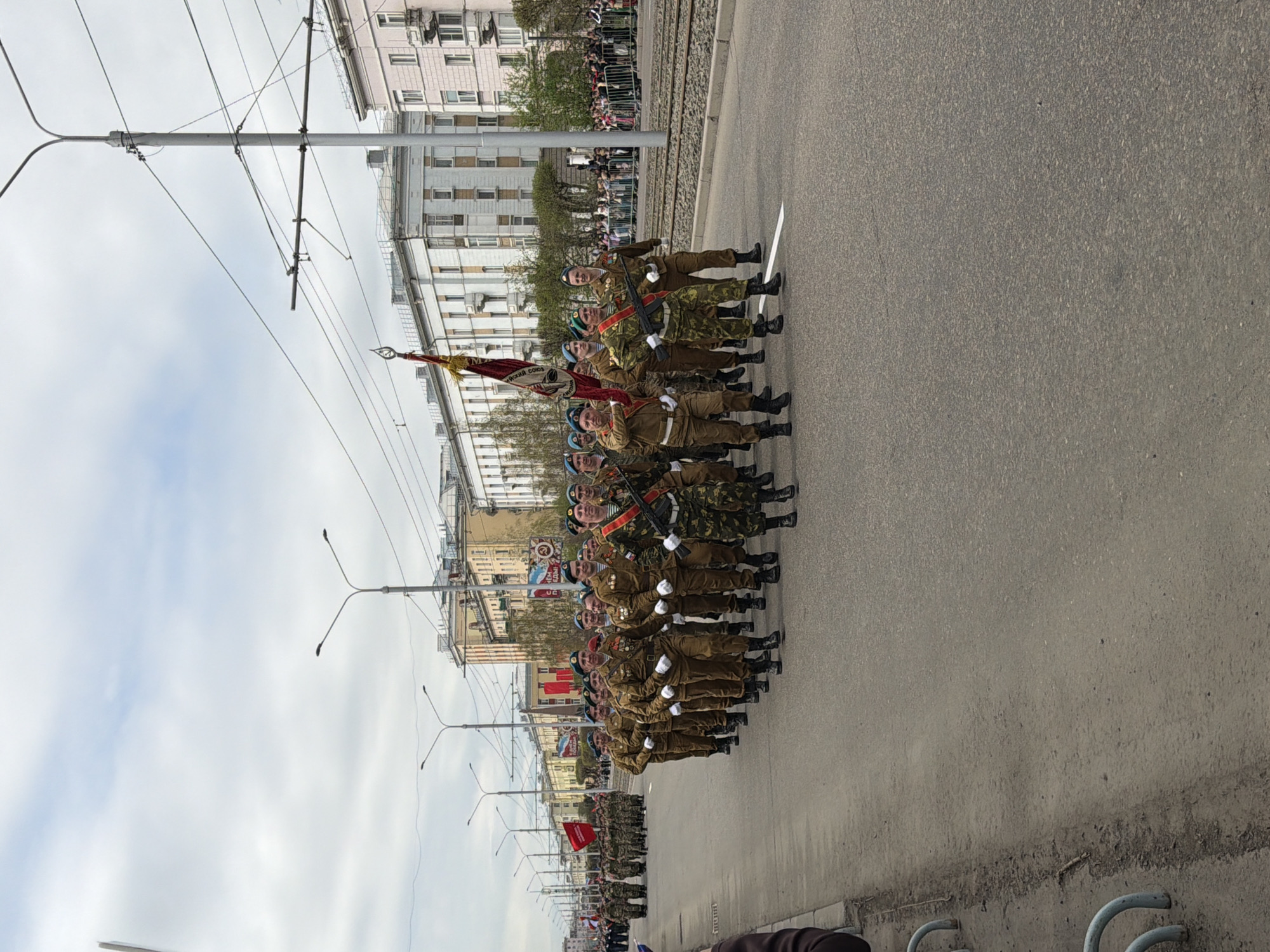 A military parade is taking place in Krasnoyarsk, Russia. A line of soldiers is marching in formation, carrying rifles and flags. The soldiers are wearing military uniforms and black boots. Some are wearing blue berets, and the flags being carried are predominantly red. They are moving along an asphalt road, with overhead power lines present. A building is on the left side of the image behind a crowd of spectators, and a railway is separating the spectators from the marching soldiers.