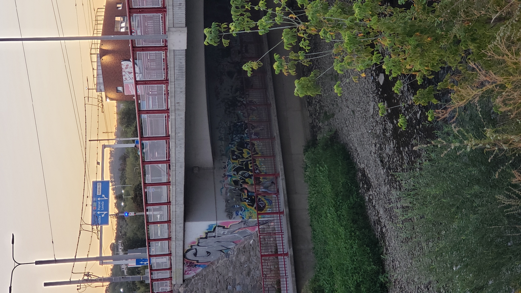 The image shows a bridge in Prešov, Slovakia. On the bridge, there is a sign pointing to Košice, Poprad, and Levoca. There is also graffiti on the wall underneath the bridge. A stream runs alongside the bridge. The sky is orange and yellow, indicating sunset. There are some trees and plants in the foreground.