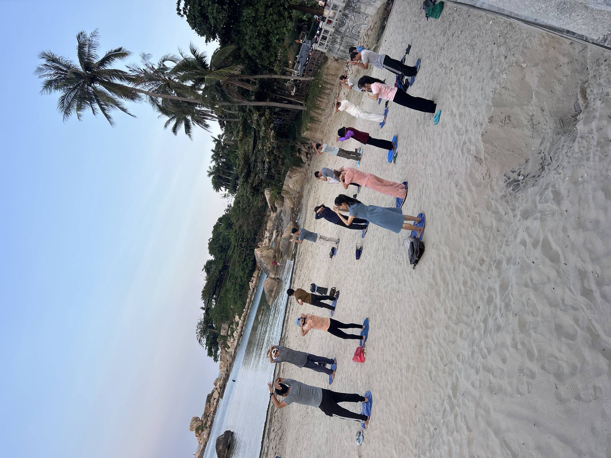 The image depicts a group of people participating in what appears to be a yoga or stretching session on a beach. They are arranged in rows on blue mats, performing a coordinated exercise with their hands raised towards their foreheads. The beach setting is a sandy area near a body of water, likely the sea. Behind the group are palm trees and other vegetation, suggesting a tropical or coastal environment. In the background, there are hints of buildings and structures, indicating the proximity to a developed area. The location is identified as Bac Nha Trang Ward, Vietnam, given the details of the city in the prompt. The overall scene suggests a calm and healthy activity in a natural setting within the coastal city.