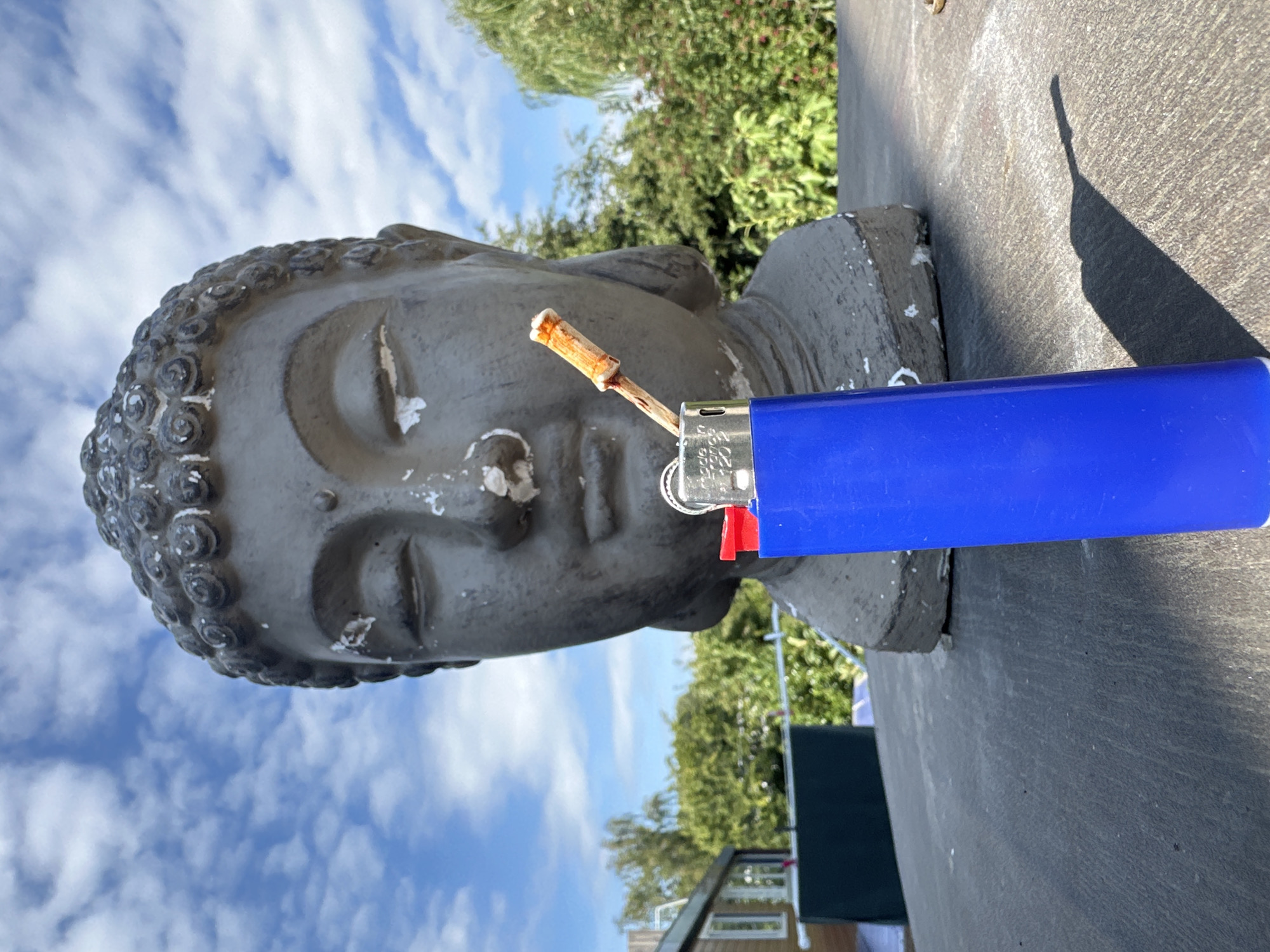 An eye-level, close-up outdoor shot features a gray statue of a Buddha's head against a bright blue sky filled with white, fluffy clouds. In the foreground, a bright blue lighter with a red igniter is positioned vertically, with a dried twig protruding from its opening, appearing as if it's being lit. The statue's face is partially obscured by the lighter, and its texture shows signs of wear and tear, with white patches and cracks. Behind the statue, lush green trees and foliage are visible, suggesting a garden or park setting. To the left, a glimpse of a building with a light-colored facade and dark windows can be seen. The ground in the foreground is a dark, textured surface, casting a distinct shadow from the lighter. The scene appears to be located in Lelystad, Netherlands, judging by the building style and vegetation.