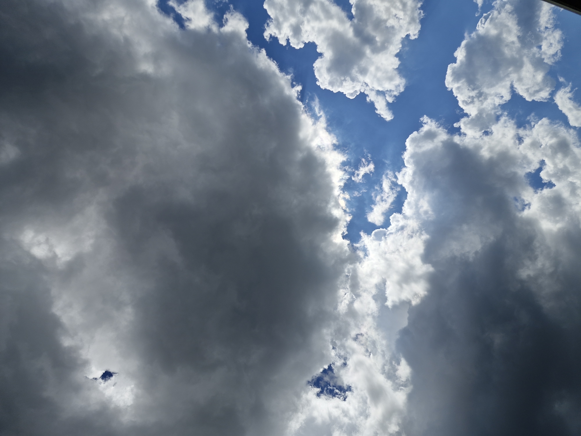 The image displays a dynamic sky scene in Unknown, Unknown, characterized by a significant contrast between dark, heavy cloud formations and areas of bright blue sky with white clouds. The upper-left quadrant and central-left area are dominated by dense, dark grey clouds, presenting a somber appearance. Within these dark clouds, in the upper-left, a small, irregular opening reveals a patch of vivid blue sky, bordered by intensely bright white cloud edges. A prominent diagonal line runs from the upper-right to the lower-middle of the image, marking the boundary where a large mass of dark grey cloud transitions into a vibrant blue sky. This blue sky section, particularly pronounced in the mid-right and lower-right, features numerous scattered, well-defined white cumulus clouds. Sunlight illuminates the edges of some darker clouds and strongly brightens the white clouds against the blue background. No ground features, man-made objects, or individuals are present.
