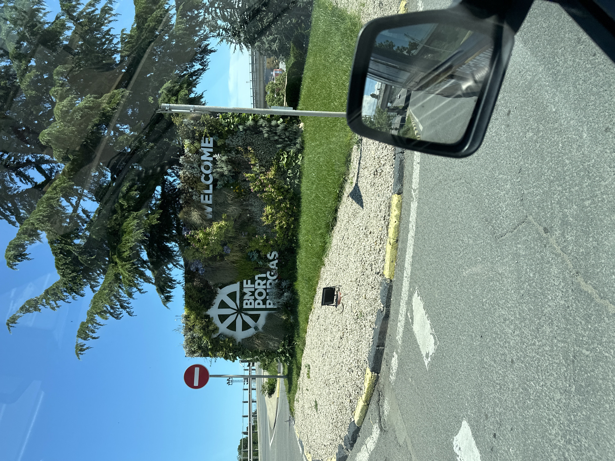 This image captures a vertical view of a roadside scene, likely taken from inside a vehicle. The primary subjects are a large, vertical green wall covered in plants, which displays the text "WELCOME BMF PORT BURGAS" in white lettering. A large, stylized wheel symbol is also part of the signage. To the left of the green wall, lush green trees with dense foliage are visible, and beyond that, a bright blue sky suggests a clear day. To the right of the green wall, a gravel-covered area with yellow and white striped curbs leads to an asphalt road. A side mirror of a vehicle is prominent in the upper right corner, reflecting a road and some distant structures. A circular red and white "no entry" sign is visible on a pole in the lower left portion of the image. The lighting and shadows suggest it is daytime. The overall impression is one of arriving at a location, possibly a port, on a sunny day.