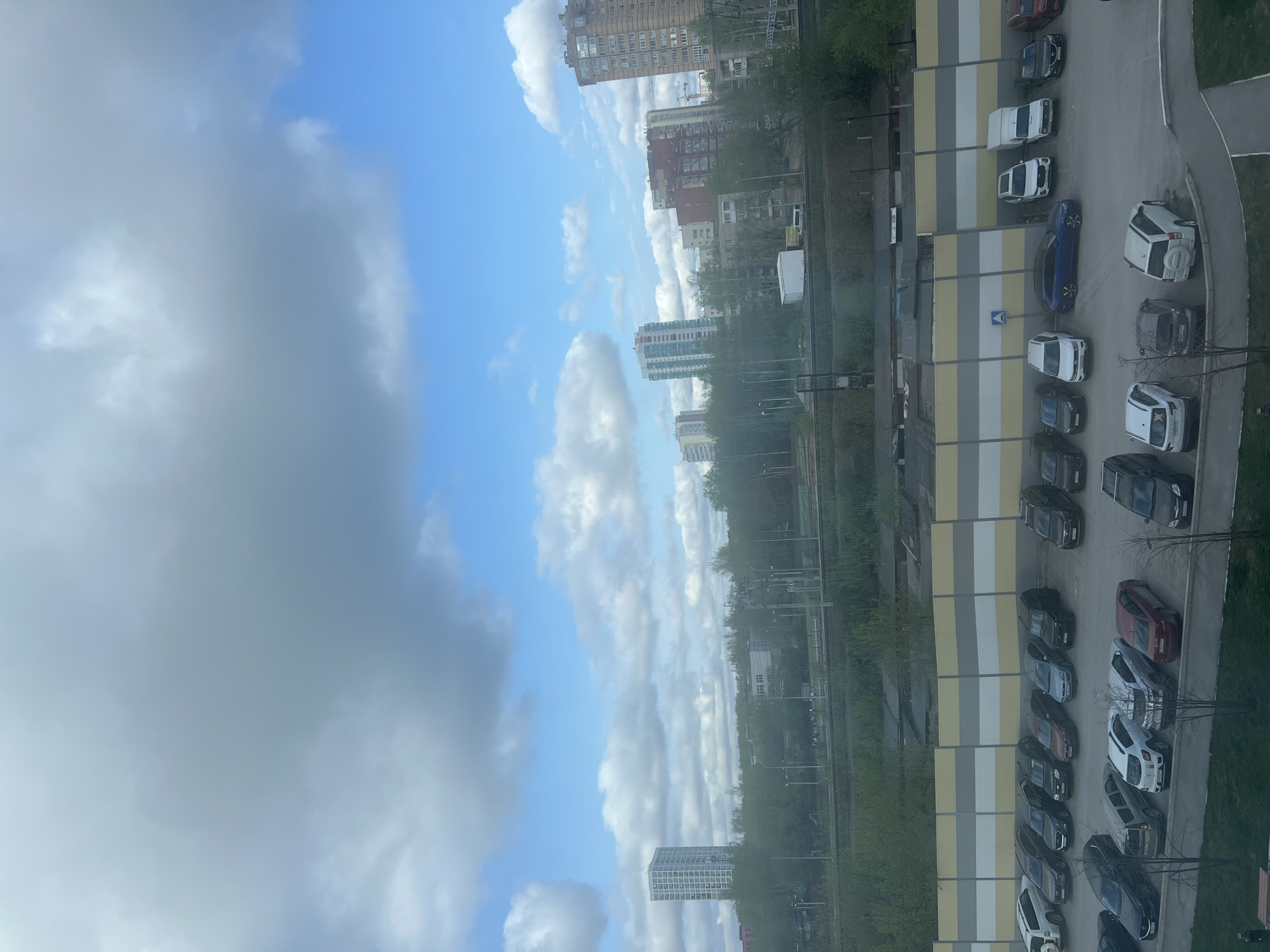 The image depicts a view from a high-rise building in Perm, Russia, looking towards the sky and a cityscape. The sky is a mix of blue and clouds. Below the clouds are residential buildings of varying heights and designs. A long, linear parking lot is visible along the right side of the frame, filled with cars parked in rows. A grassy area with trees is seen between the buildings and the parking lot. The scene appears to be taken during daylight.