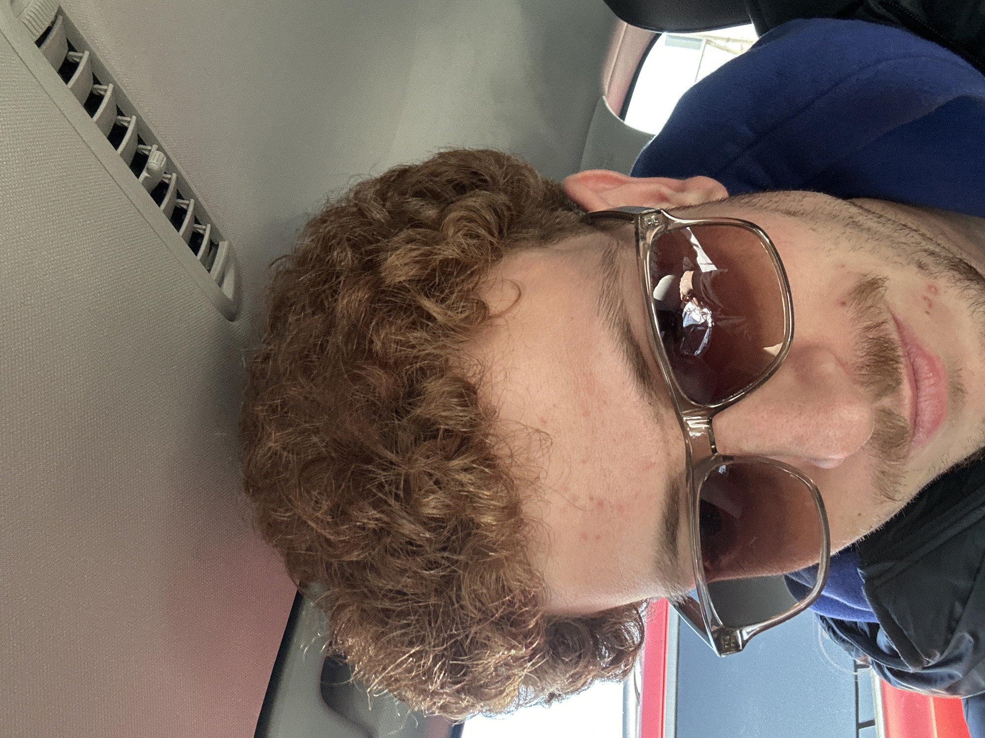 A person with curly, reddish-brown hair is wearing sunglasses, a blue sweatshirt, and a black jacket. The person appears to be inside a vehicle, as indicated by the car's interior features such as the air vent and ceiling panel. The person is likely in New York City given the red and white background visible through the car window is the color of a typical bus in New York City.