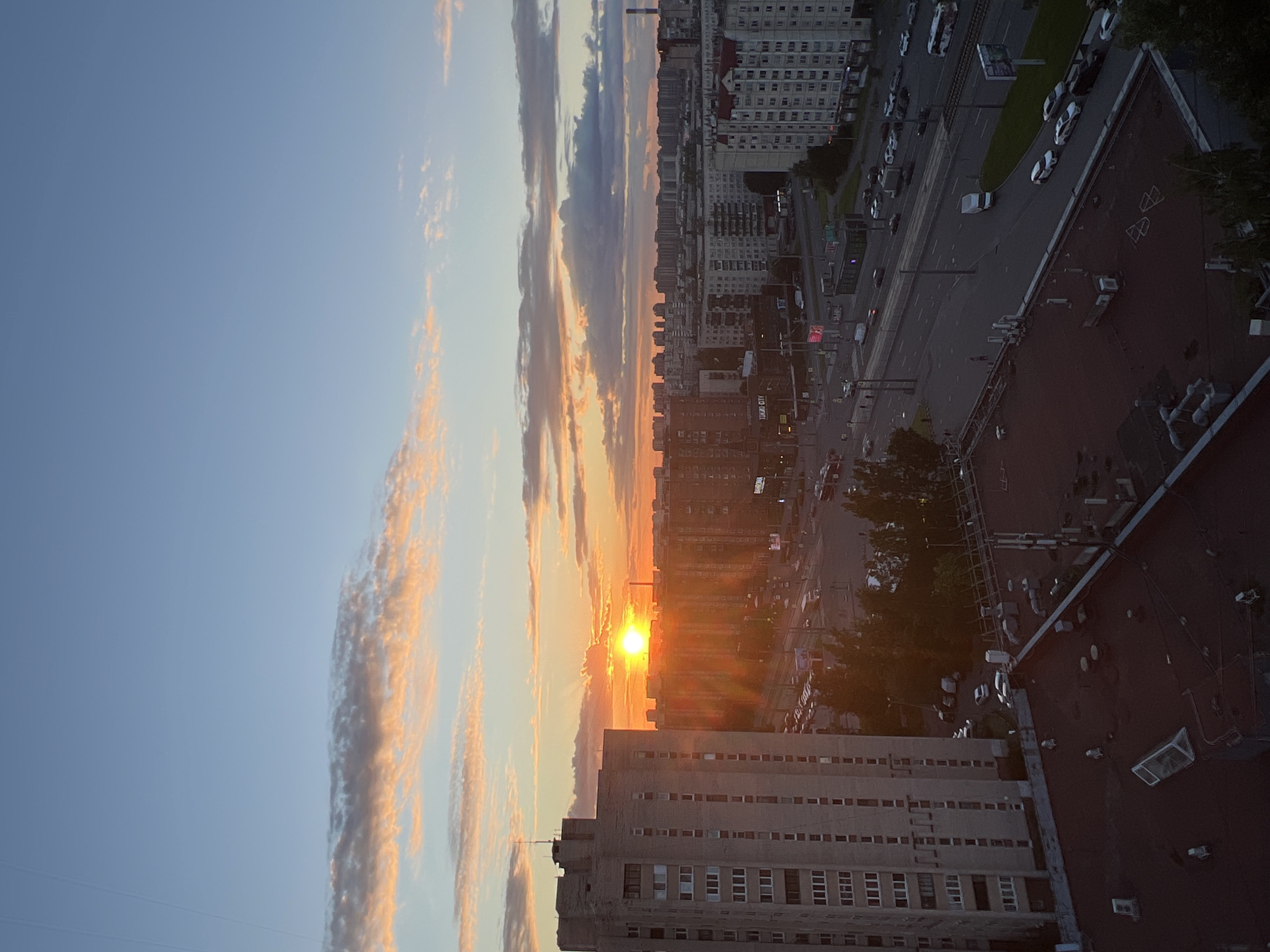 This media file captures an urban scene at sunset. The sky is a vibrant mix of blue and orange, with streaks of clouds illuminated by the setting sun. The sun itself is a bright, low-hanging orb, casting long shadows and creating a warm, golden glow.
The foreground is dominated by the rooftops of several buildings. One building in particular, made of concrete and featuring numerous windows, is prominently visible. Its scale suggests it's a multi-story residential or office building. Further back, the cityscape unfolds with more buildings, some appearing taller and more modern with white facades, while others are older, darker structures.
A street runs through the scene, lined with cars and trees. The presence of traffic indicates the city is still active despite the approaching evening. The road appears to be a major artery, possibly with tram lines visible in some areas. The overall impression is of a bustling urban environment at the end of the day.
Notable details include the distinct lines of windows on the buildings and the variety of vehicles on the street. The time of day is clearly sunset, suggested by the dramatic sky and lighting. The weather appears clear, with the sunset being the dominant atmospheric feature. While no specific people are clearly discernible, the presence of cars and the overall urban setting imply human activity. There is a small amount of text visible on a sign on a building, but it is too small and blurry to be read. The scene evokes a sense of peace and transition, as day turns into night.