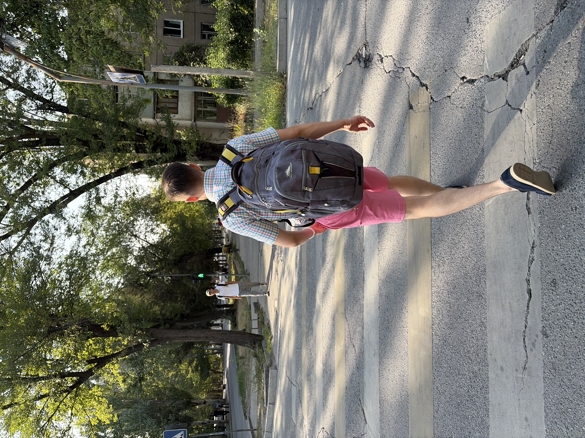 A person is crossing a street at a crosswalk, walking away from the camera. They are wearing a plaid shirt, pink shorts, and dark shoes, and carrying a grey backpack with yellow accents. Another person is visible in the background, walking on the sidewalk. The street has several cracks in the pavement. Trees line the street, and buildings can be seen in the distance. The scene appears to be in a city during daylight, with sunlight casting shadows.
