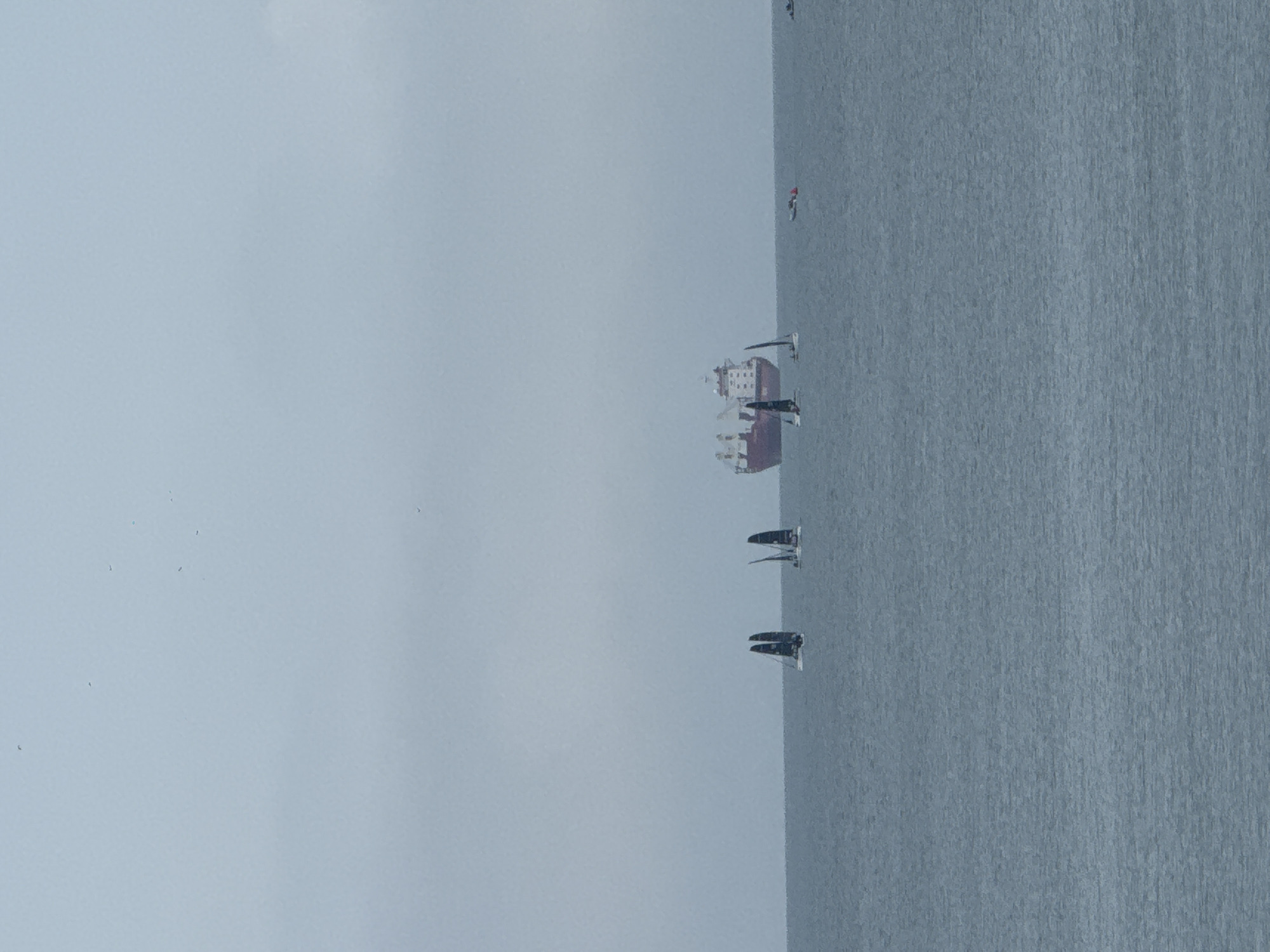 The image shows a body of water, likely the sea, with several sailboats and a large cargo ship in the distance. The sky is overcast, suggesting a cloudy day. The location is potentially near Vladivostok, Russia, considering the presence of maritime activity.