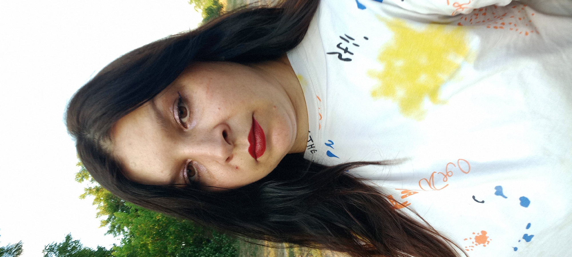 A woman with dark hair and fair skin is pictured in a close-up, outdoor shot. She is wearing a white t-shirt with colorful abstract patterns and text. Her expression is somber, with her gaze directed slightly downwards. The background consists of greenery and a bright sky. The image does not provide any information that would allow for a determination of its location within the city of Klyntsi, Ukraine, or any other specific location.