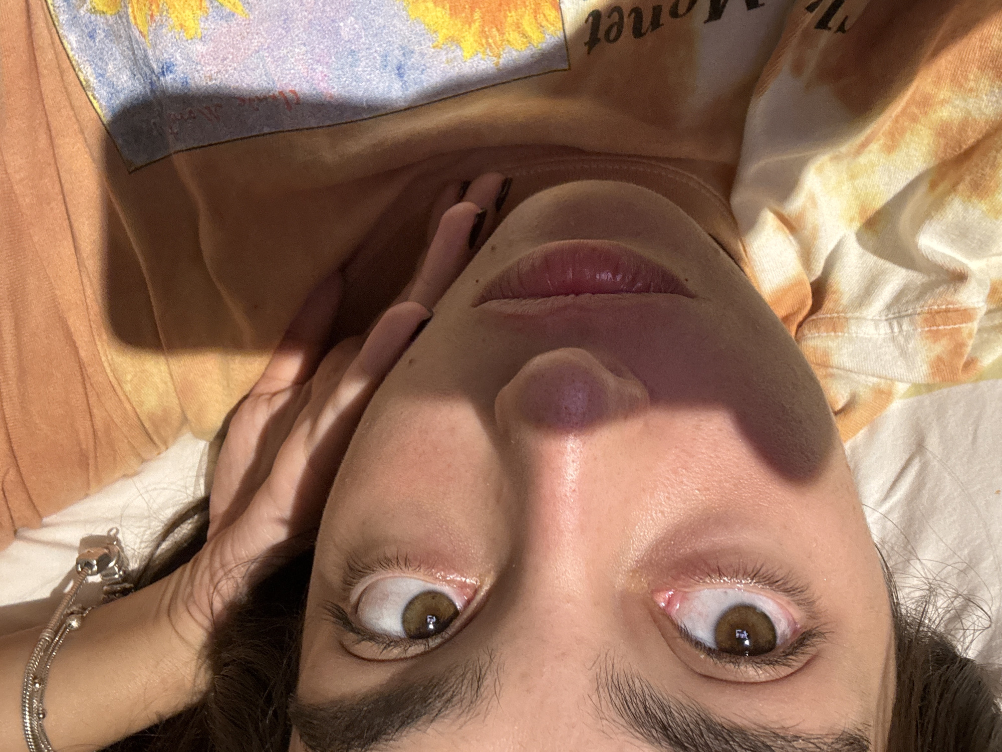A close-up, tilted view of a person's face looking upwards. The person has fair skin, brown eyes, dark eyebrows, and slightly parted lips. A hand with painted black nails rests near the left side of the face. The person is wearing a t-shirt with a tie-dye pattern in shades of orange, yellow, and white. The image is lit by sunlight, creating shadows across the face. A silver bracelet is visible on the person's left wrist. The location is unknown and not discernable from this image.