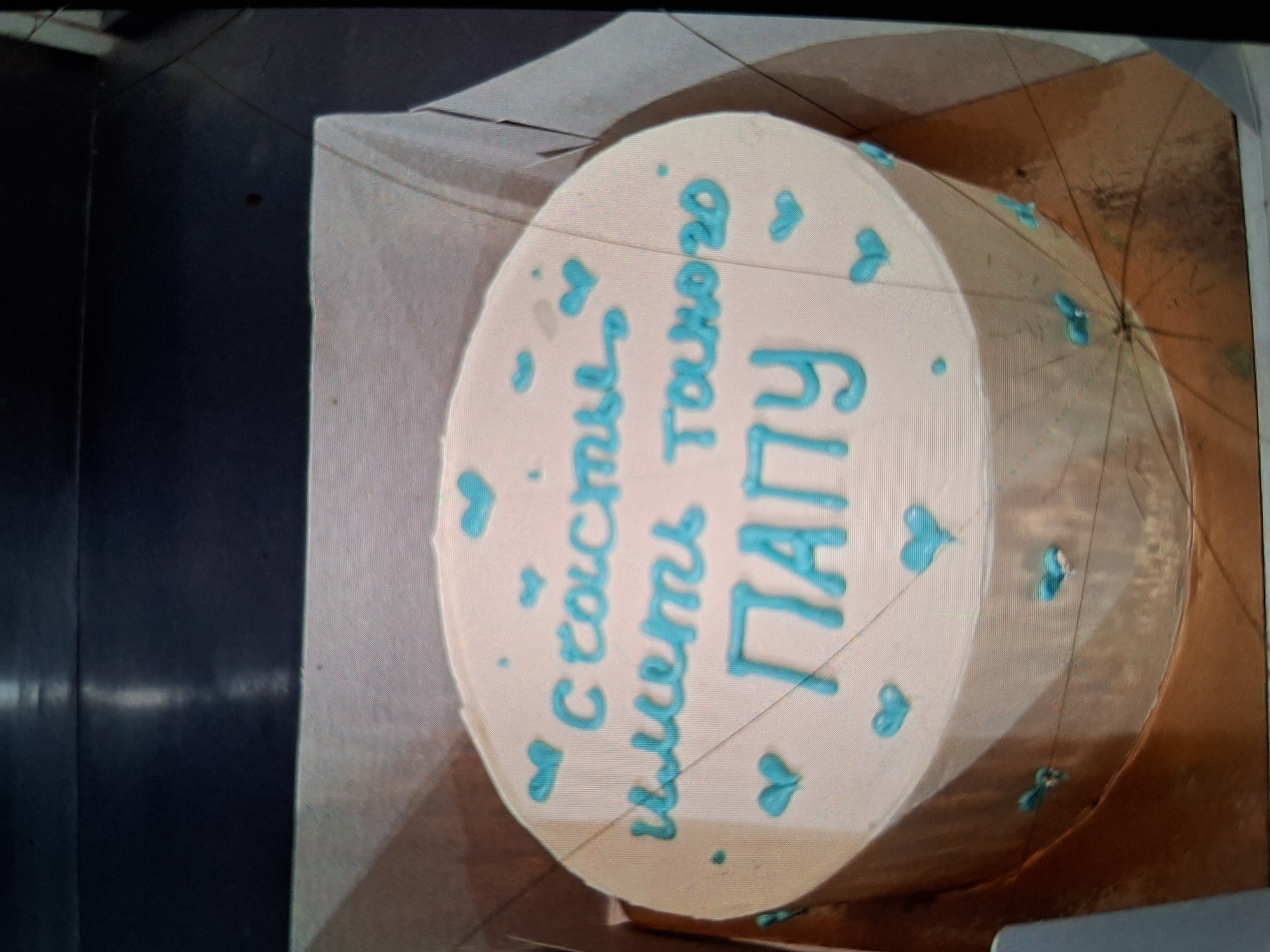 The image shows a round cake decorated with blue frosting. The cake is placed on a gold circular board. The cake has a light pink frosting and is adorned with blue icing details in the shape of hearts and dots. Text in blue icing is written on the cake in Russian. The text reads "Счастья иметь такого ПАПУ" which translates to "Happiness to have such a DAD". The scene is indoors, with the cake on a table. The lighting is artificial, suggesting an indoor event or preparation. The focus is on the cake itself, highlighting the decorative elements and the inscribed message.
