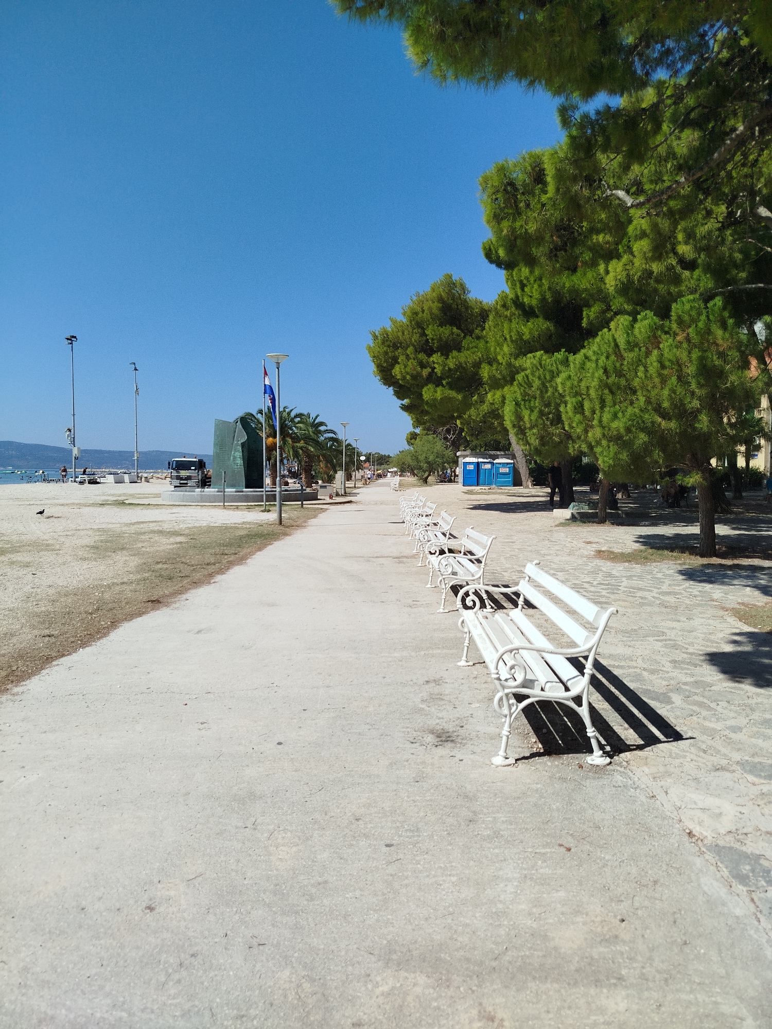 The image depicts a paved walkway along a sandy beach on a clear, sunny day. A row of white benches lines the walkway, casting shadows on the ground. Beyond the walkway, a sandy beach stretches towards the water, where several boats are visible. A memorial statue with a flag and palm trees stands on the left side of the walkway. In the distance, hills and a clear blue sky form the backdrop. Several blue portable toilets are located further down the beach, near a line of trees. The scene suggests a public recreational area, likely in a coastal city. Specifically, the memorial statue is known as the Monument to the Partisan Defenders of Omiš.