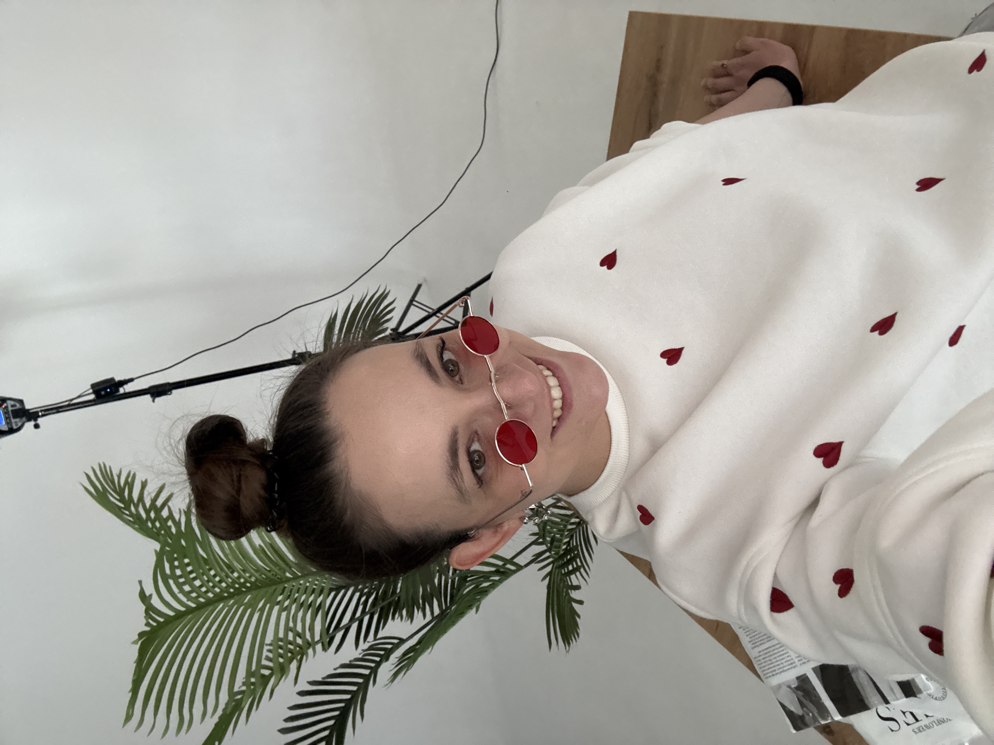 A woman smiles at the camera while wearing red, round sunglasses. She is wearing a white sweatshirt with small, embroidered red hearts. Her hair is pulled back into a bun. Behind her are a large artificial palm plant and a white wall. A lighting rig is visible in the top left of the image. A wooden table or surface is visible on the right. A label for the brand "S&R JONES LOVERS" is partially visible in the lower right corner. There is no definitive evidence to place the location in the city of Chaikovsky, Russia.