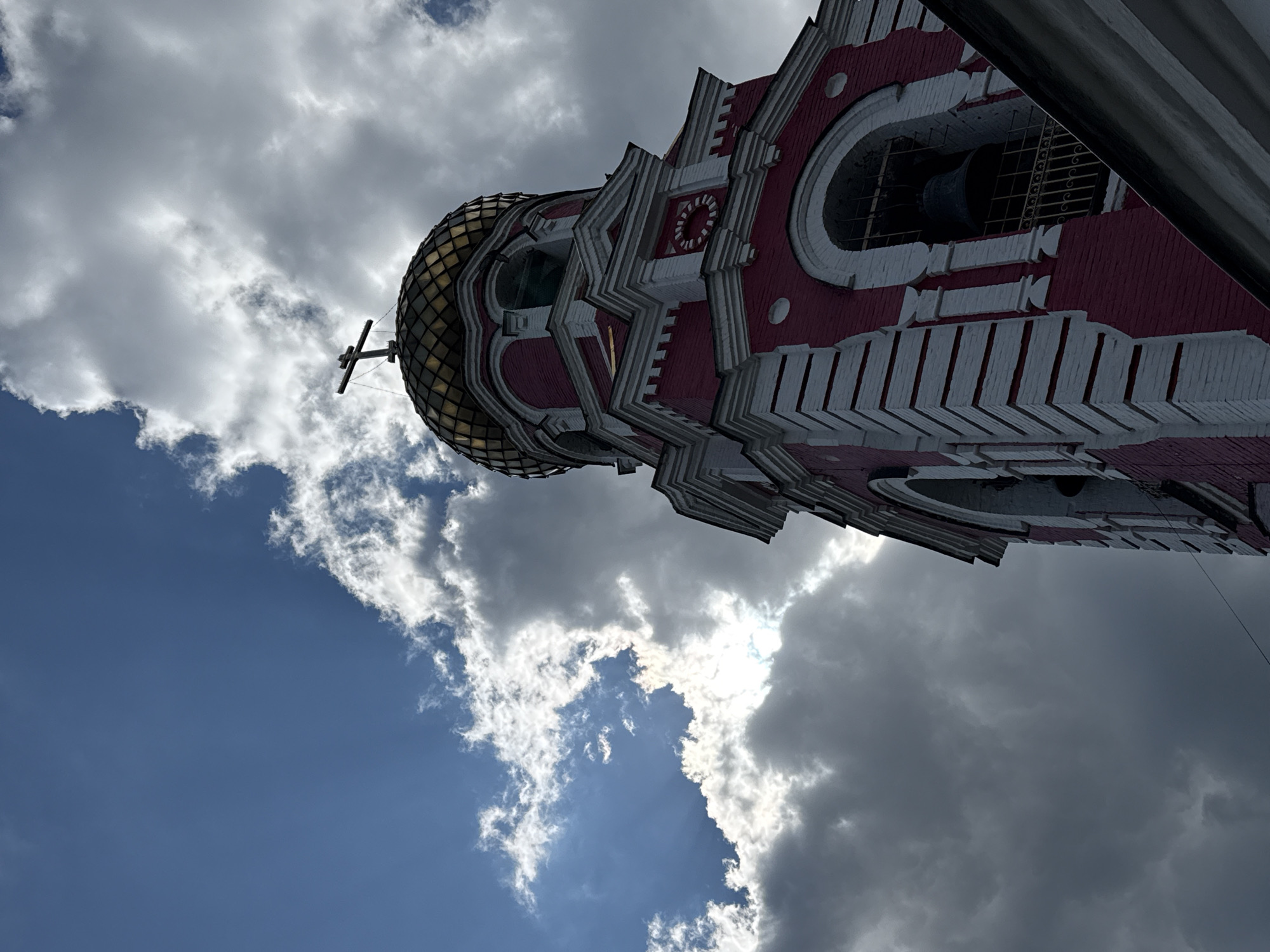 An upward-angled shot shows the red and white bell tower of the Church of the Entry of the Lord into Jerusalem in Dmitrov, Russia. The tower has a gold, onion-shaped dome topped with a cross. The tower's architectural details are intricate with repeating patterns. A large bell is visible in one of the tower's arched openings. The sky is partially overcast with both blue and gray clouds.