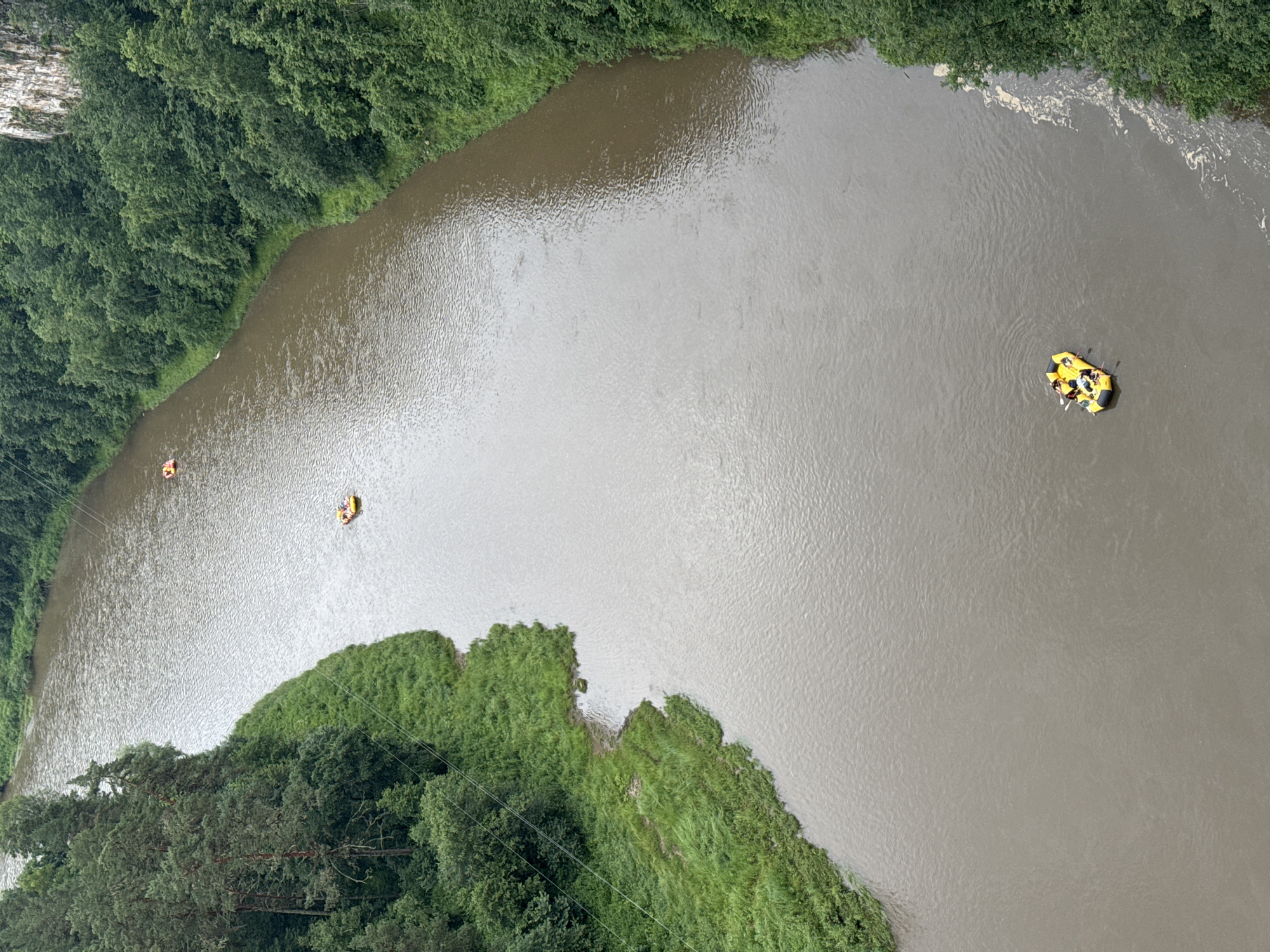The image shows an aerial view of a wide, murky river bordered by lush green trees and vegetation. The river is not perfectly still; there are ripples and some swirling patterns on its surface, suggesting a gentle current. Along the banks, dense green foliage covers the land, with some areas of lighter green grass leading down to the water's edge.
In the river, there are two inflatable rafts with people on them. One raft, closer to the left bank, is yellow and orange and appears to have at least one person in it, who is wearing an orange life jacket. Further out in the river, a second, larger yellow raft is visible with multiple people inside. They are paddling, indicating that they are engaged in a recreational activity like rafting or kayaking.
The overall impression is of an outdoor recreational scene in a natural environment. The lighting suggests it is daytime, possibly overcast given the muted light and lack of strong shadows. The dense greenery implies it is summer or late spring. There is no visible text in the image. The location context provided is Kul'metovo, Russia.