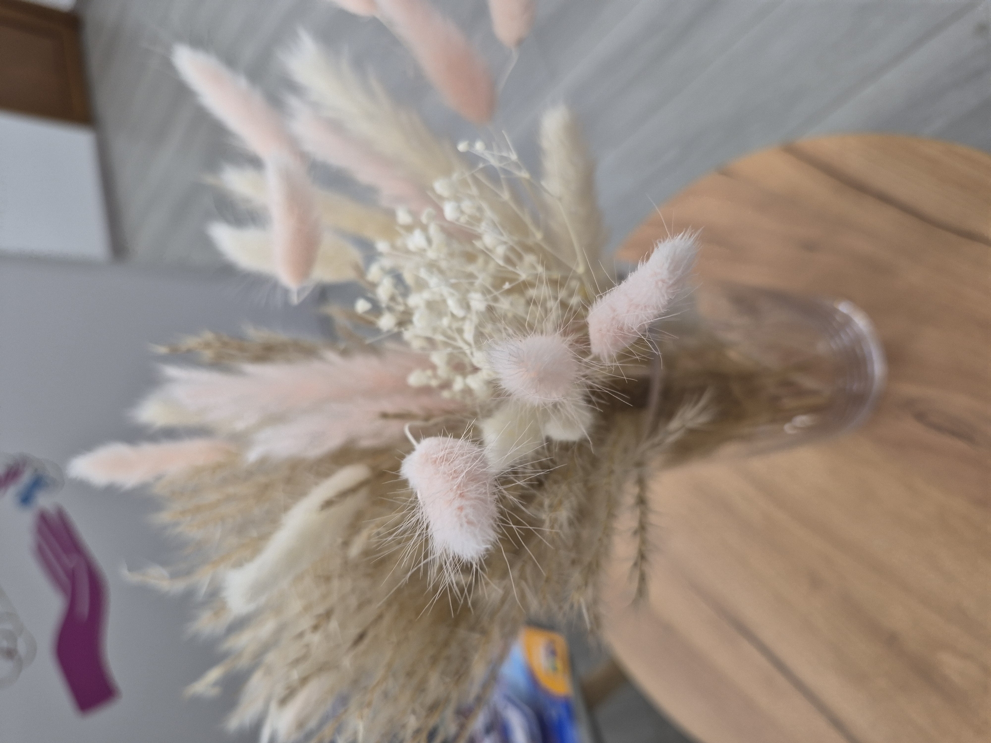 An overhead view captures a decorative bouquet of dried flowers and grasses arranged in a clear glass vase, placed on a light brown wooden table. The bouquet features a mix of textures and subtle colors, primarily consisting of light beige and brown feathery pampas grass-like elements. Interspersed among these are delicate clusters of small white dried flowers, possibly baby's breath, and distinctive fluffy, soft pink/peach-colored bunny tail grass (lagurus), which adds a vibrant yet muted accent.
The wooden table displays a natural grain and a smooth finish. The background behind the arrangement is a blurred light gray surface, likely a wall or paneling, exhibiting faint linear patterns. In the soft-focus areas, other objects are visible: a glimpse of a brown structure, a white rectangular item, and a noticeable magenta-colored hand shape or cutout on the left. Below the bouquet, a blurred object with blue and yellow tones can be partially seen. The scene is lit with soft, diffused light, typical of an indoor setting. No specific time of day or outdoor weather conditions are discernible. No visible text is present in the image. The setting is in Yerevan, Armenia.