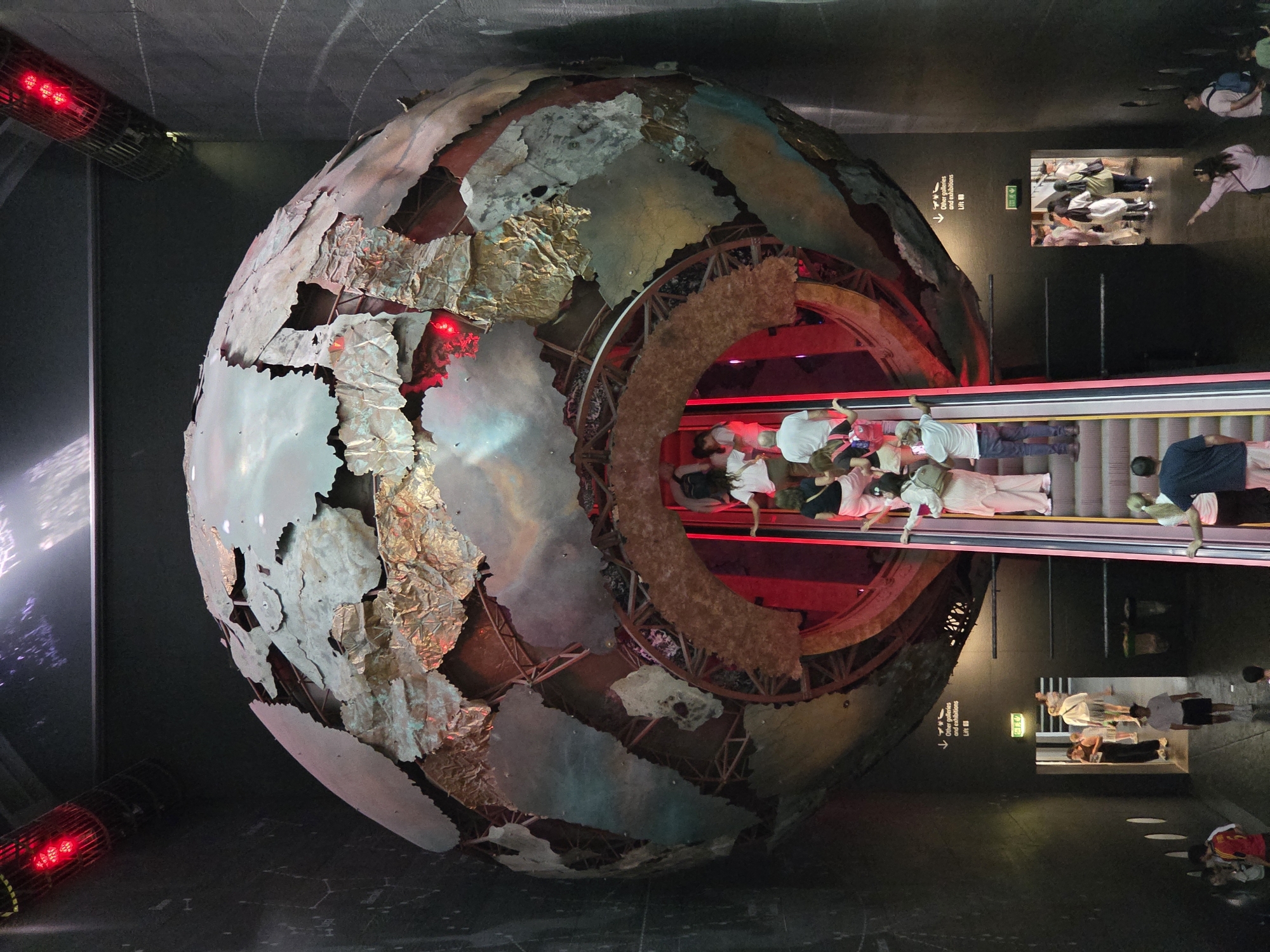The image shows a large, deconstructed globe-like structure, possibly an art installation or exhibit, viewed from above. An escalator passes through the center of the structure, carrying people. The globe's exterior is made of metal pieces resembling landmasses, some appearing weathered or torn. Red lighting is visible inside the structure and also from a vent on the side. Some people are walking along the side. A sign points to other galleries and exhibitions. The image does not include any information that would allow it to be placed within the city of London, United Kingdom.