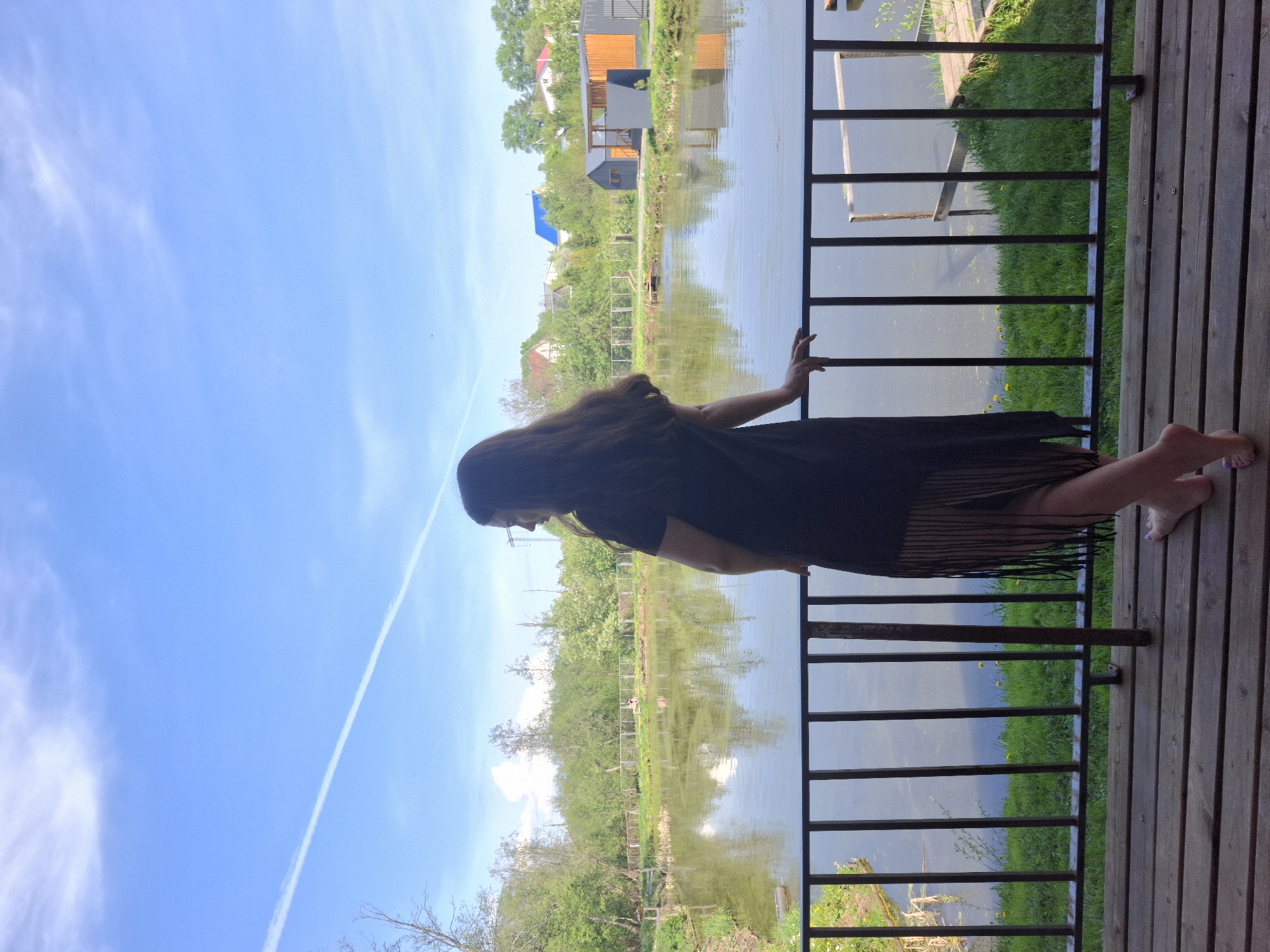 A vertical, full shot captures a woman on a wooden deck, seemingly overlooking a body of water.
The woman has long, dark hair, and she is clad in a dark fringed garment. She is leaning forward, resting her hands on the railing. Her feet are bare.
The deck is constructed of wooden planks, and a dark railing stands between the woman and the water. Beyond the water is a green landscape with trees and houses, with a clear blue sky above.
The geographical location is Malyi Bukor, Russia.