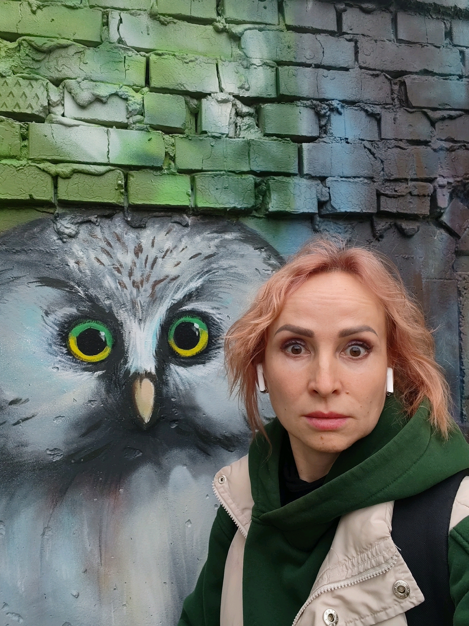 This is a medium shot, eye-level, outdoor photograph of a woman standing in front of a graffiti mural in Chaikovsky, Russia. The woman has light pink hair, brown eyes, and is wearing a green hooded sweatshirt under a cream-colored jacket. She has white wireless earbuds in her ears and is looking directly at the camera with a surprised expression. The graffiti mural features a large, gray owl with bright green and yellow eyes. The background is a brick wall, with the lower portion covered in the owl mural and the upper portion painted in green and blue, with some areas of exposed brick. The lighting suggests it is daytime.