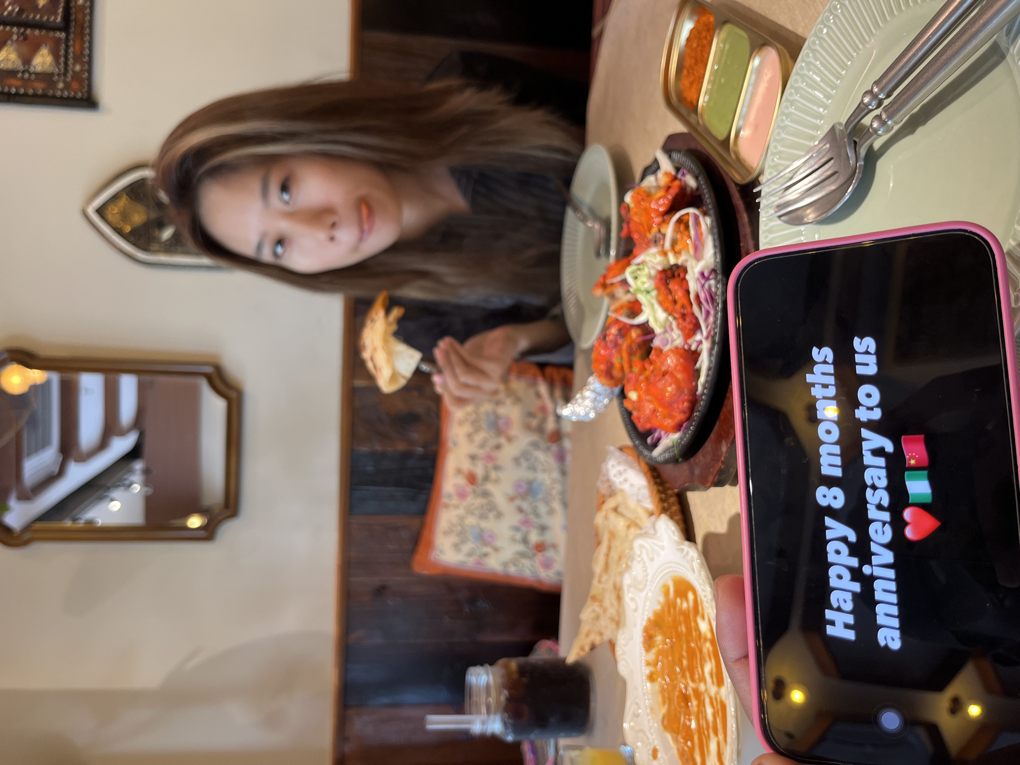 The image captures an indoor scene at what appears to be an Indian or South Asian restaurant. A young woman with long brown hair, wearing a dark top, is seated at a wooden table, looking towards the camera. She is holding a piece of flatbread (naan or roti) with a fork, suggesting she is in the middle of a meal.
In front of her, on the table, are several dishes. A prominent one is a sizzling plate of tandoori chicken or a similar red-marinated meat dish, garnished with shredded cabbage or salad. Beside it, there's a light green/mint-colored plate with a fork and spoon resting on it. Small metal condiment dishes hold orange, green, and pink sauces or chutneys. Another plate displays flatbread accompanied by a yellow-orange dip or sauce. A mason jar with a straw contains a dark beverage, possibly cola. The table is adorned with a patterned fabric cushion or placemat.
In the foreground, a person is holding a pink-cased smartphone, facing the camera. The phone screen displays text that reads: "Happy 8 months anniversary to us" followed by emojis of the Chinese flag, the Nigerian flag, and a red heart. This suggests a celebration or special occasion for a couple.
The background features warm, dim lighting, decorative framed mirrors, and ornate wall hangings, contributing to an inviting restaurant ambiance. The time of day appears to be evening. The woman's expression is neutral, looking directly at the camera. The location context is Yuzhong District, China.