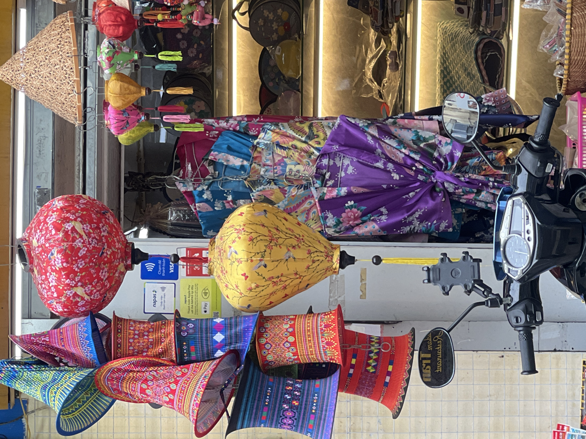 This image depicts a street scene in Hanoi, Vietnam, likely near a shop selling traditional Vietnamese items. In the foreground, a black scooter is parked, with its handlebars and a portion of the dashboard visible. A phone mount is attached to the handlebars. The background features a storefront with various items displayed, including colorful conical hats, lanterns, and patterned robes. Several payment logos are visible on the shop's facade, indicating acceptance of digital payments. The overall atmosphere suggests a vibrant marketplace.