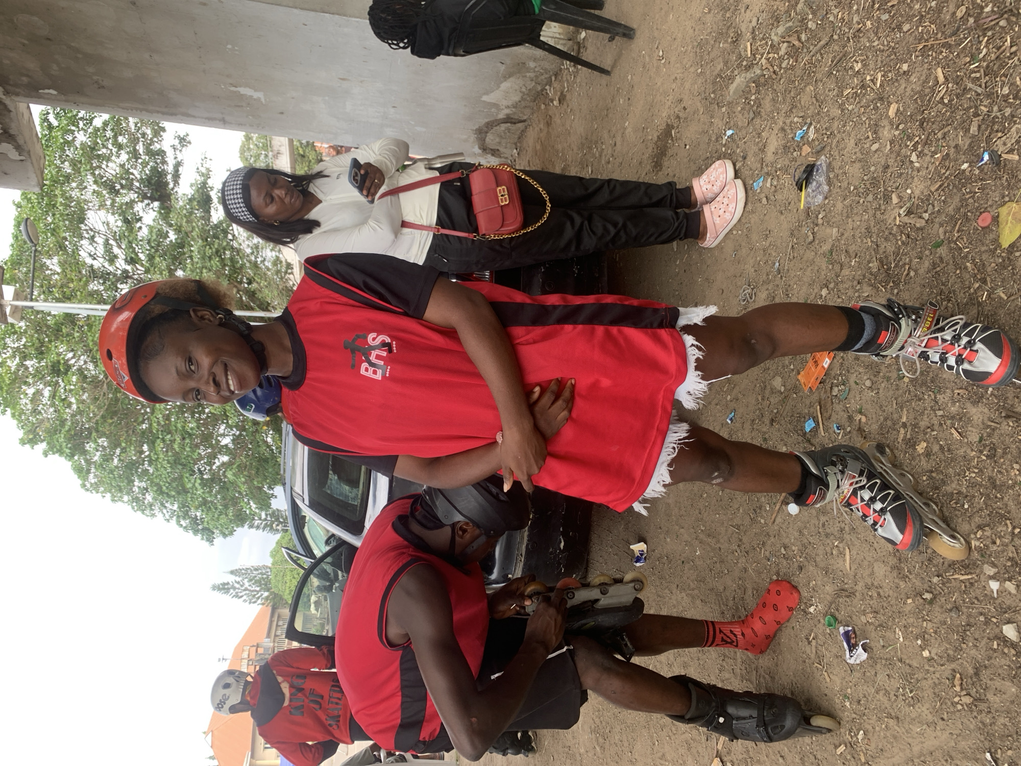 In Alagbede, Nigeria, a group of young people are gathered near a parked vehicle. A woman in the foreground, wearing a red and black sleeveless jersey and a red helmet, smiles at the camera. She is wearing rollerblades and has white trim on her shorts. To her left, a man in a red shirt and helmet bends over to adjust his rollerblades. He is wearing black shorts and red socks. Behind them, another woman in a white jacket and black pants looks at her phone, holding a red purse with a gold chain. The ground is dirt and littered with debris. Trees and a building are visible in the background.