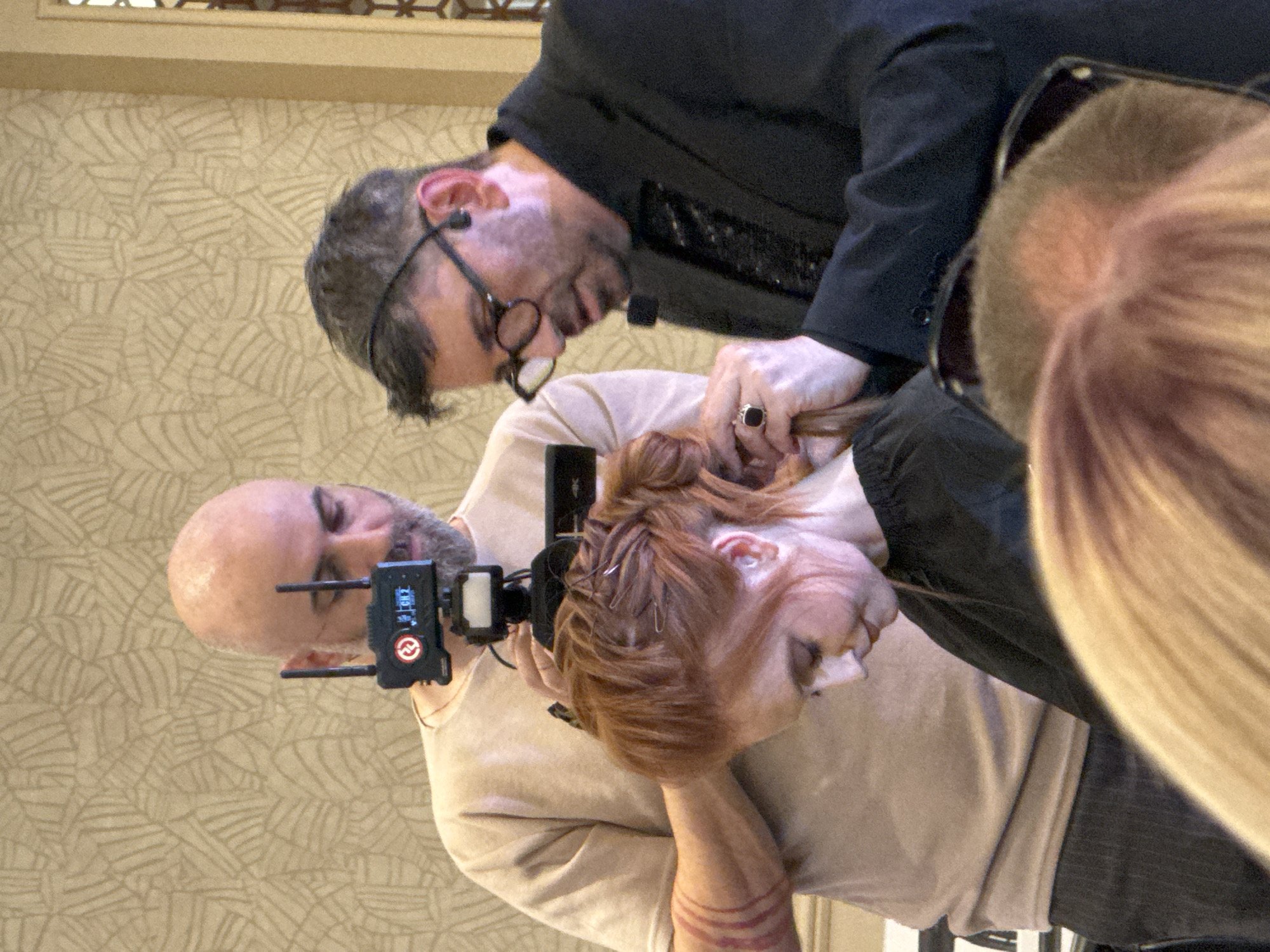 Two men are working on the hair of a woman with red hair. One man, wearing a black blazer and glasses, is holding the woman's hair. The second man is bald and is wearing a beige shirt and has a black device attached to his head. The background is a patterned wall. The location and city are unknown.