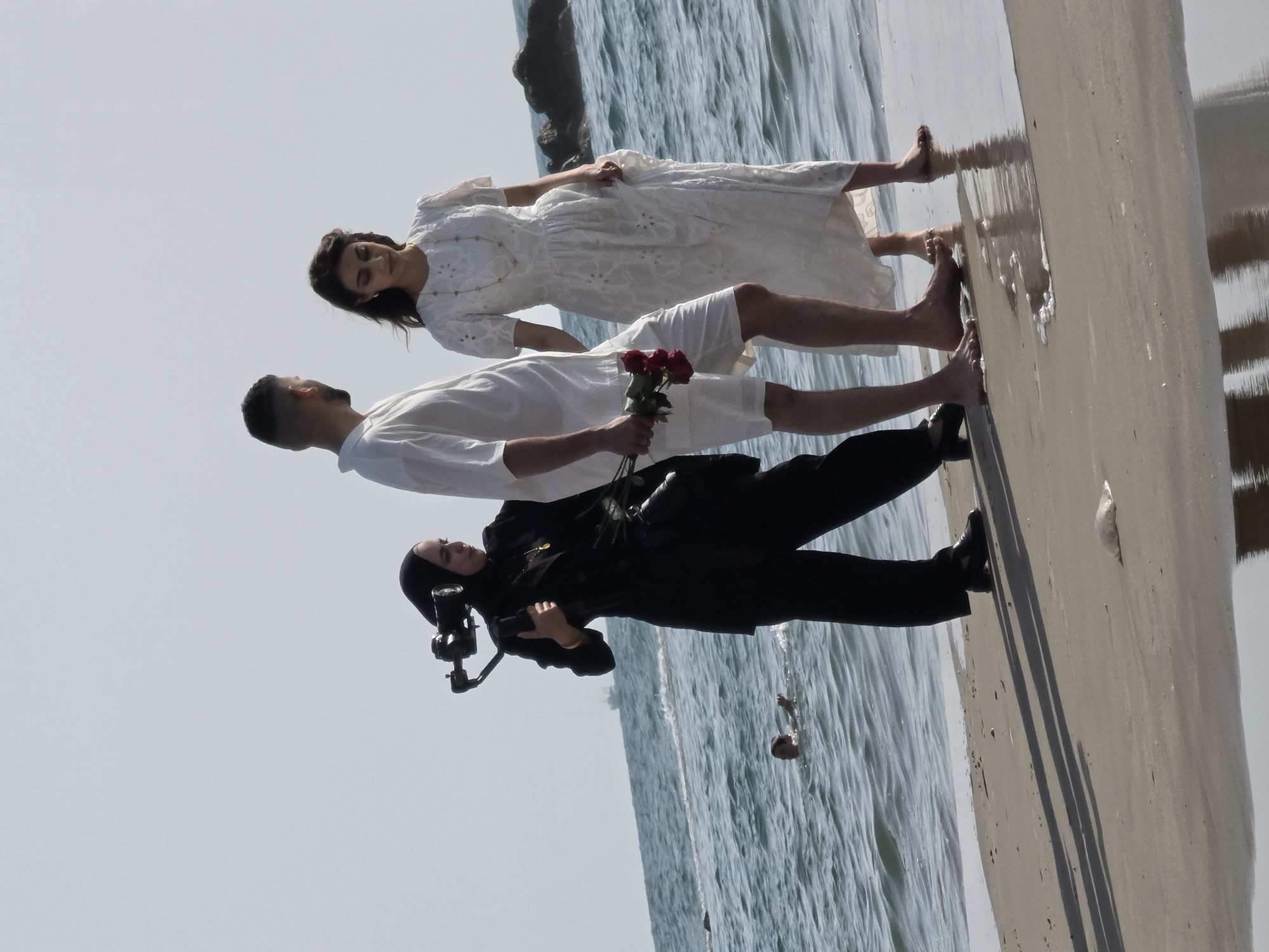The photograph depicts a photoshoot taking place on the beach in Bat Yam, Israel. A man in white shorts and shirt is lying on the wet sand, holding a bouquet of red roses. A woman in a white dress is lying next to him. A photographer, wearing a black hijab and pantsuit, is kneeling to take pictures. The scene is set against the backdrop of the sea and sandy beach.