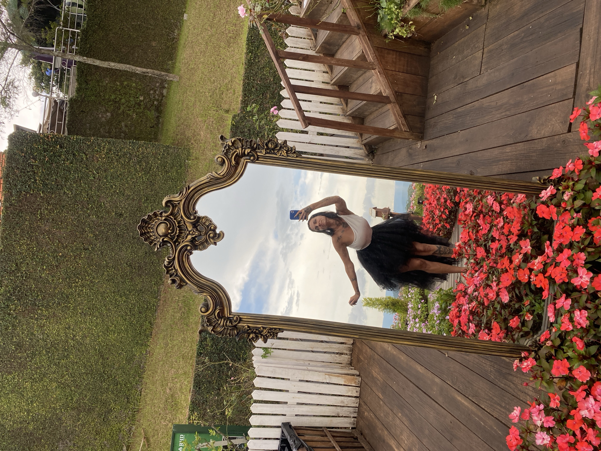 A woman is taking a selfie in a large, ornate, gold-framed mirror. She is wearing a white top and a black tutu, and appears to be smiling. The mirror reflects the sky and clouds, as well as a garden filled with red and pink flowers. The setting seems to be outdoors, with a wooden deck and a white picket fence visible in the reflection. There is also a wooden staircase leading up to a higher level. The woman's legs are visible in the reflection, and she has tattoos on them. The overall mood is playful and cheerful. The image appears to have been taken during the daytime, with natural light illuminating the scene. There is a small green sign with white text at the bottom of the image, but the text is not legible. The location is described as Da Lat, Vietnam.