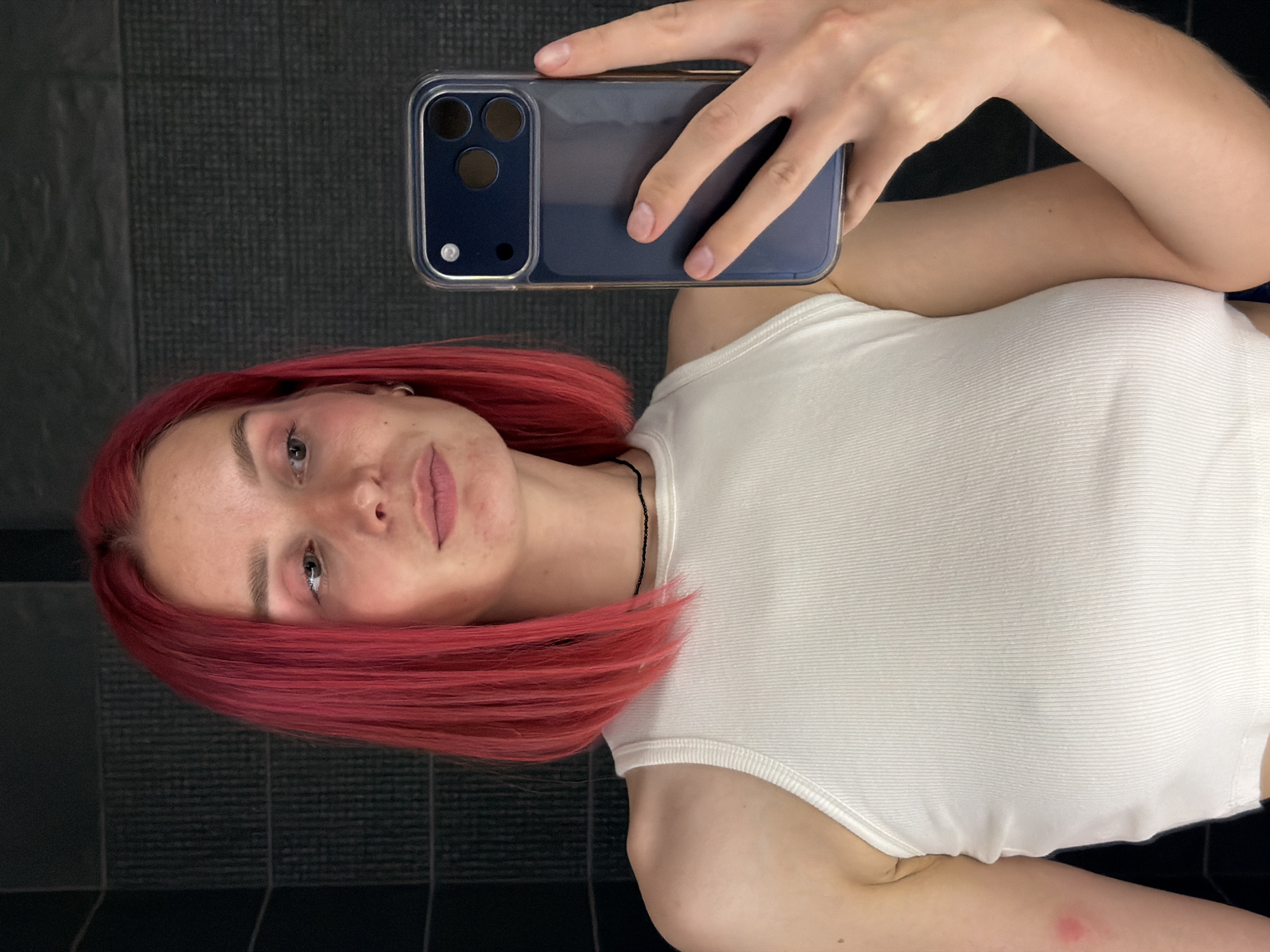 In a dimly lit bathroom with dark, textured walls, a young woman with bright pink bobbed hair takes a mirror selfie. She is wearing a white ribbed tank top and a thin black choker necklace. She holds a dark blue smartphone in her right hand, angled to capture her reflection. Her left arm rests by her side, revealing a reddish mark on her upper arm. Her face is turned slightly to the left, with her gaze directed at her phone screen. The lighting is soft, highlighting her facial features and the texture of her hair and clothing. The environment appears to be a private residence in Chaikovsky, Russia.