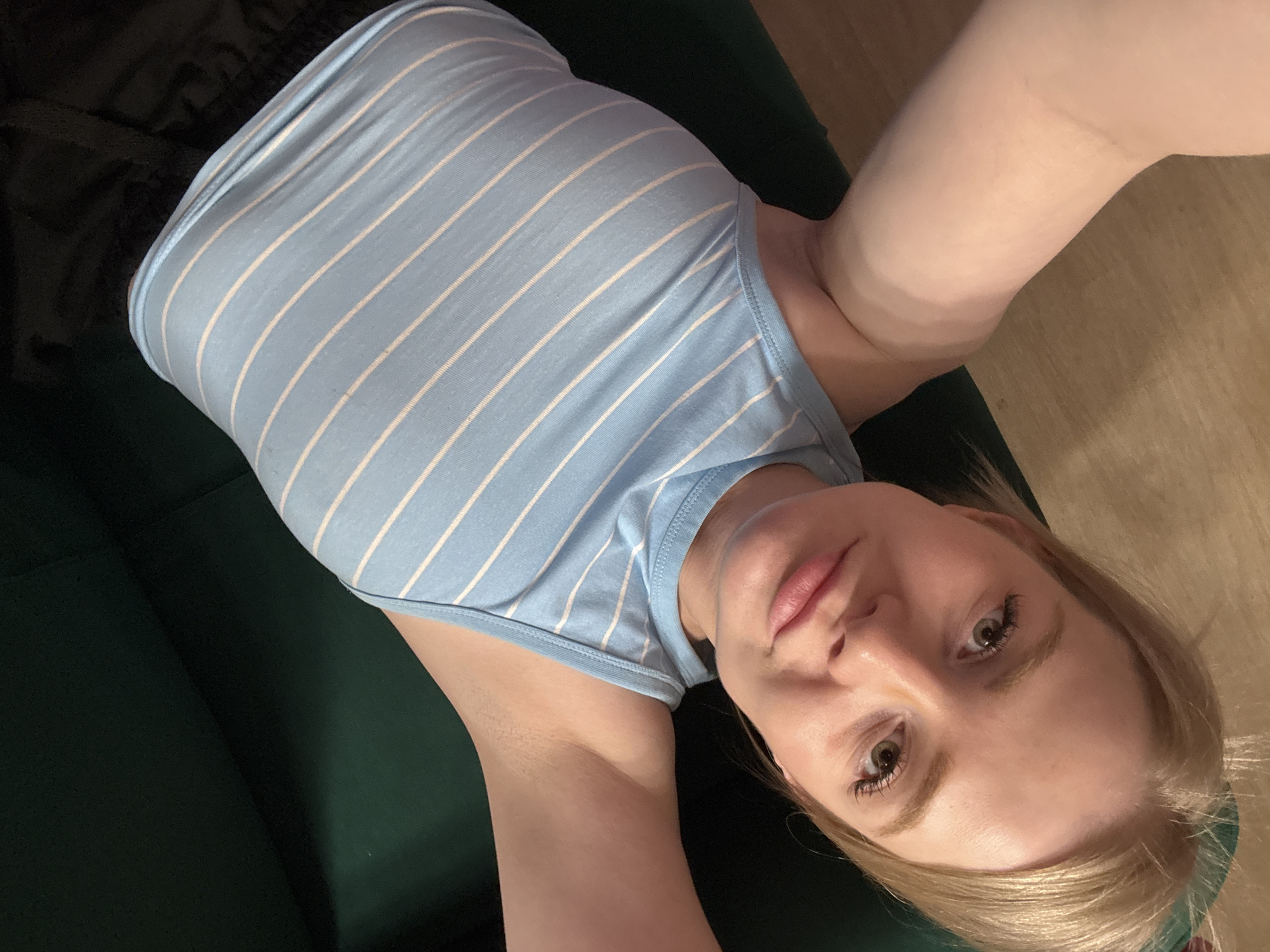 A fair-skinned person is lying on a dark green sofa, angled towards the camera. They are wearing a light blue tank top with white horizontal stripes, and black shorts are visible. The person's face is close to the camera, showing light blonde hair, green eyes, and pink lips. The background includes a wooden floor. The image does not contain enough information to determine the location in Saint Petersburg, Russia.