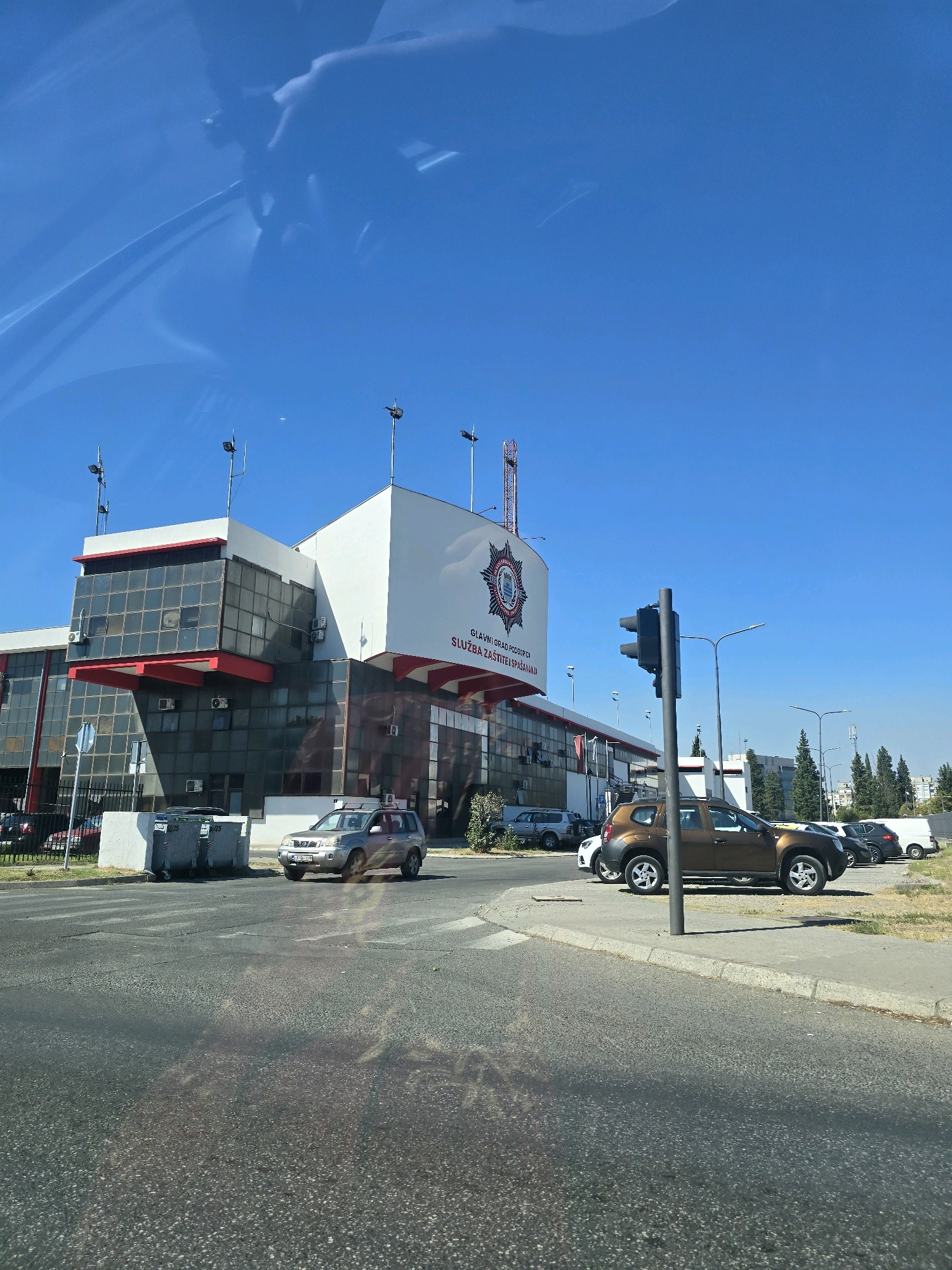 A daytime, medium-shot, exterior view captures the Podgorica Fire Department building, situated on a clear, sunny day. The building, predominantly white with red accents, features a modern design with large glass windows. In front of the building, several vehicles are parked. A silver SUV is on the road, turning right at an intersection controlled by a traffic light. A brown SUV is parked on the side of the road. The environment appears to be an urban setting, with trees and other buildings visible in the background. The sky is a vibrant blue with minimal cloud cover. The image is captured from a moving vehicle, indicated by the slight blur and reflections on the windshield.