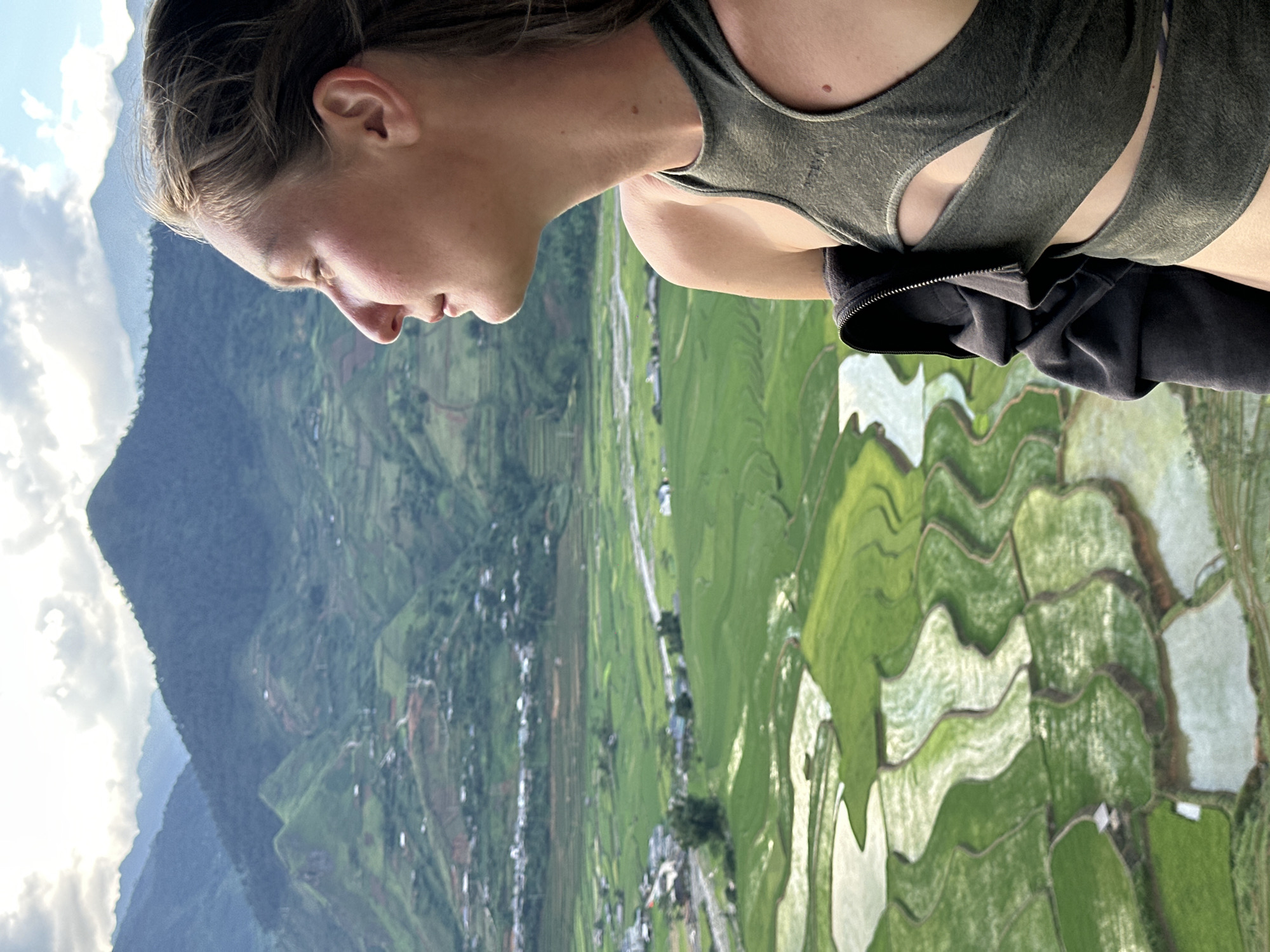 The image shows a woman in profile, positioned on the right side of the frame, overlooking a mountainous landscape with terraced rice fields in Tú Lệ, Vietnam. She is observed from the chest up, facing left, with her head slightly tilted back and her eyes closed or directed upwards. Her expression appears serene. Her hair, light brown or blonde, shows movement consistent with wind. She wears a dark olive green, high-necked athletic top featuring two horizontal cut-outs across the upper torso. A dark grey hooded jacket is draped over her left shoulder and arm. A small, dark nevus is visible on her upper right chest. The background consists of extensive, undulating terraced rice fields, predominantly green, with sections reflecting water as lighter, irregularly shaped patches. A river or stream flows through the lower valley, adjacent to a cluster of buildings forming a small settlement. Beyond the valley, multiple layers of densely forested mountains extend into the distance. The sky above is partly cloudy, displaying both white clouds and areas of blue, with bright illumination from the sun.