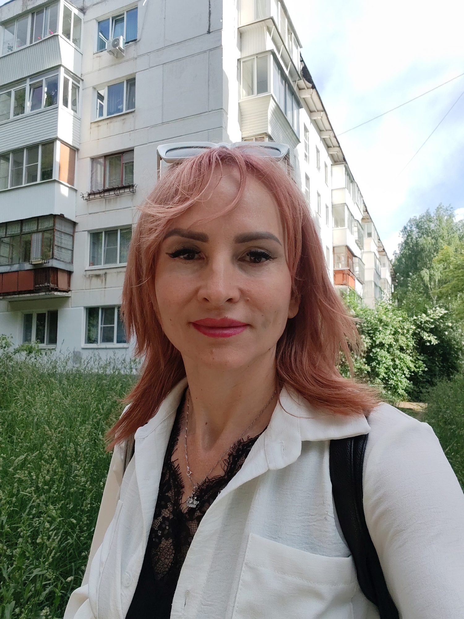 A woman with pink hair is taking a selfie outdoors, in front of a white apartment building. She is wearing a white button-down shirt over a black lace top, and a silver necklace with a small pendant. Her sunglasses are resting on her head. The building appears to be in a residential area, with greenery and trees surrounding it. The sky is visible in the background, with some clouds. The woman is looking directly at the camera.